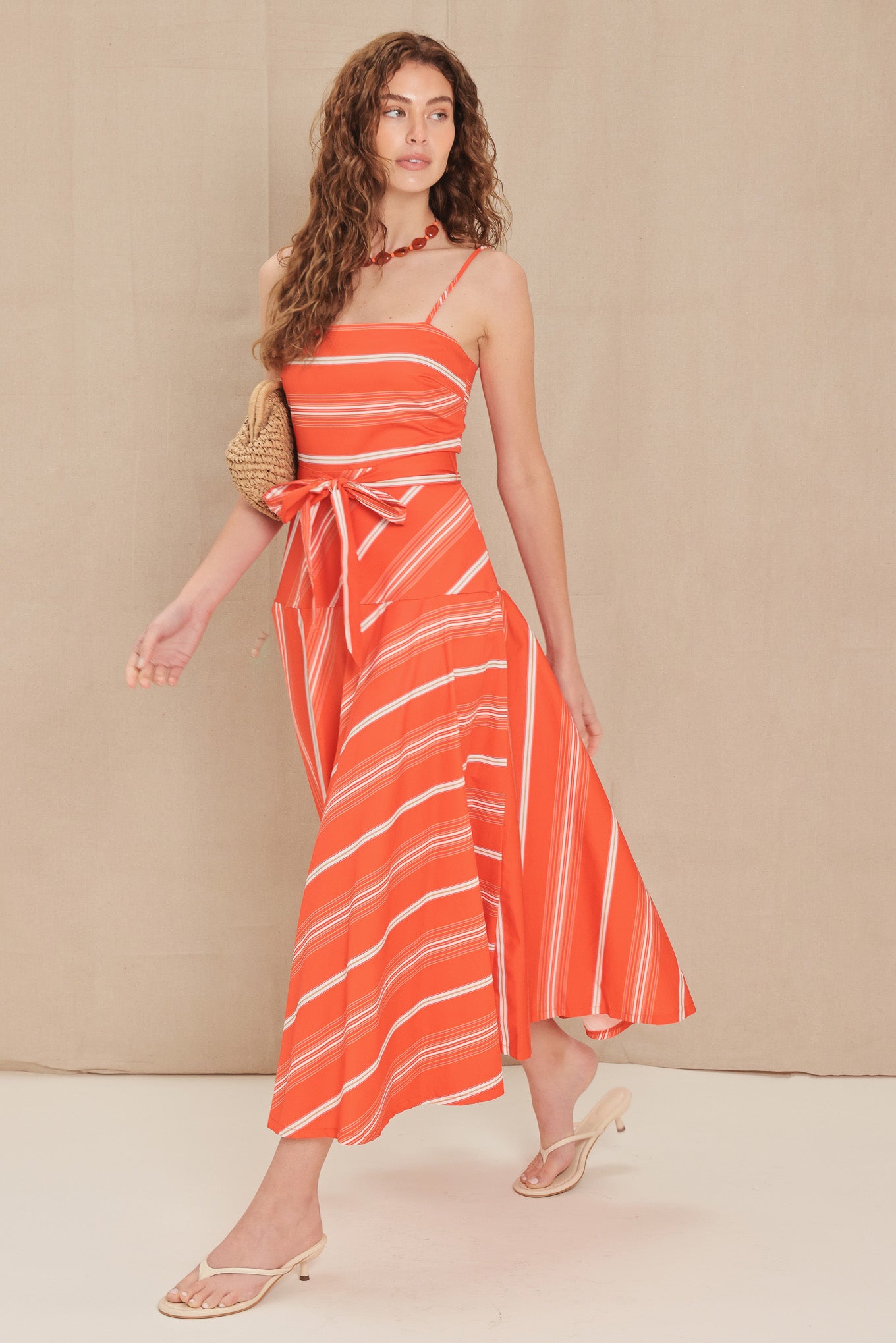 A woman models a bright orange and white striped midi dress with thin straps, a tie waist, and a flowing skirt, paired with a woven clutch and neutral sandals against a beige studio backdrop.