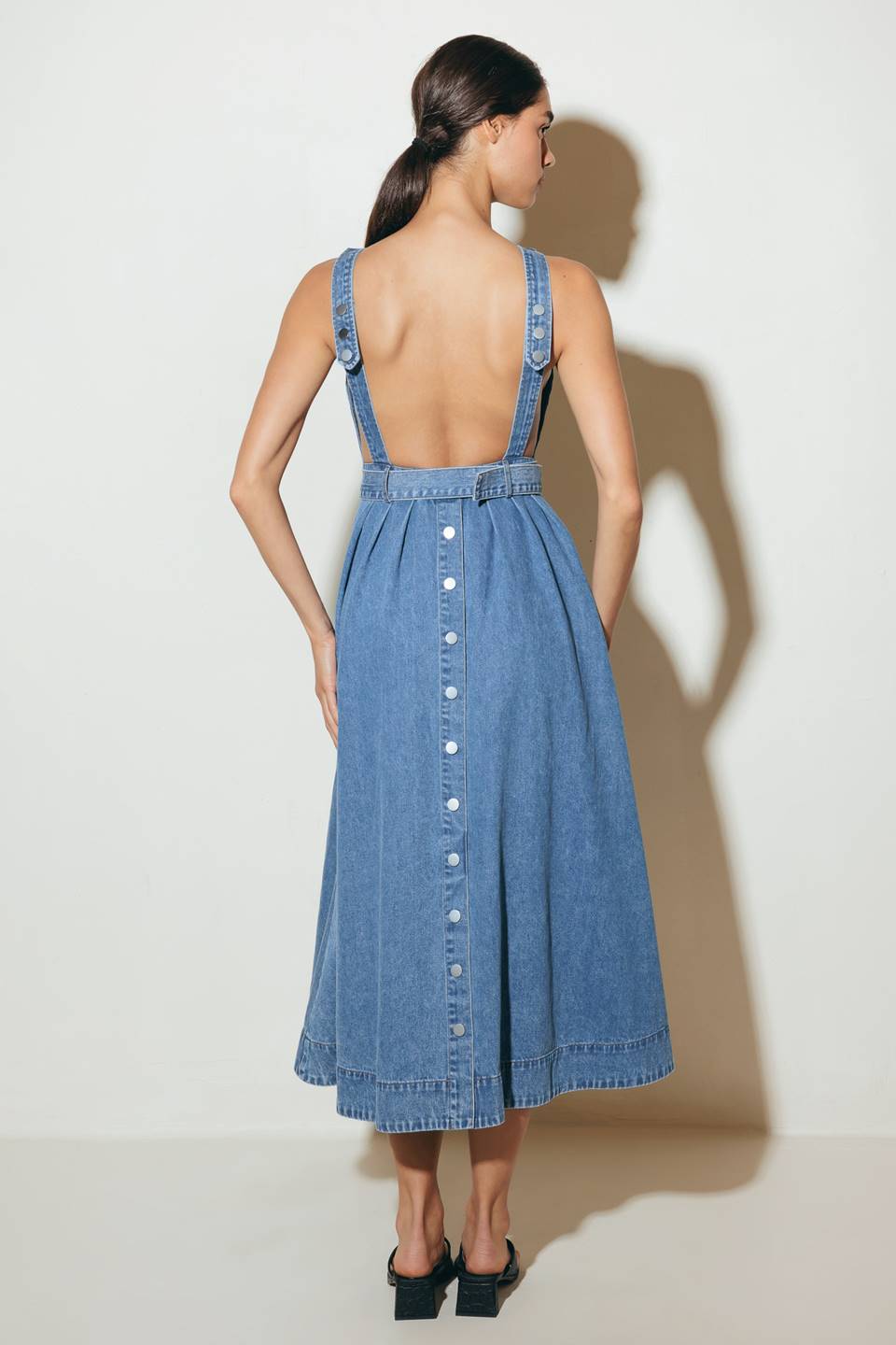 A washed denim midi dress featuring boat neckline, sleeveless, self belt, full skirt, side pockets, bare back, adjustable strap and back button down closure