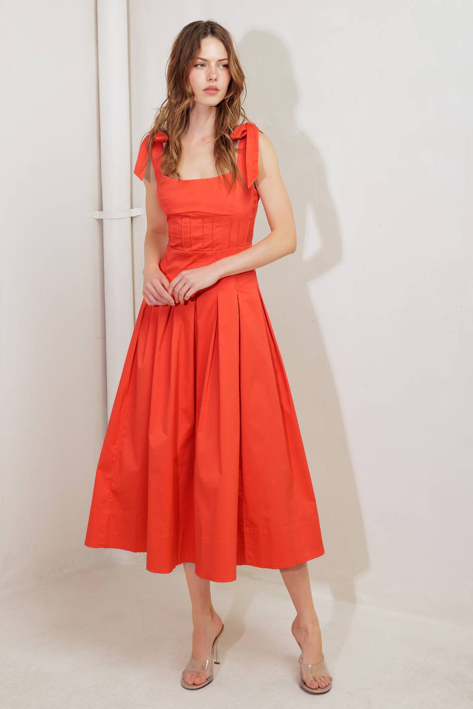 A solid red woven midi dress featuring straight neckline, shoulder ties, pintuck waist, inverted pleated skirt and bare back with straps and zipper closure