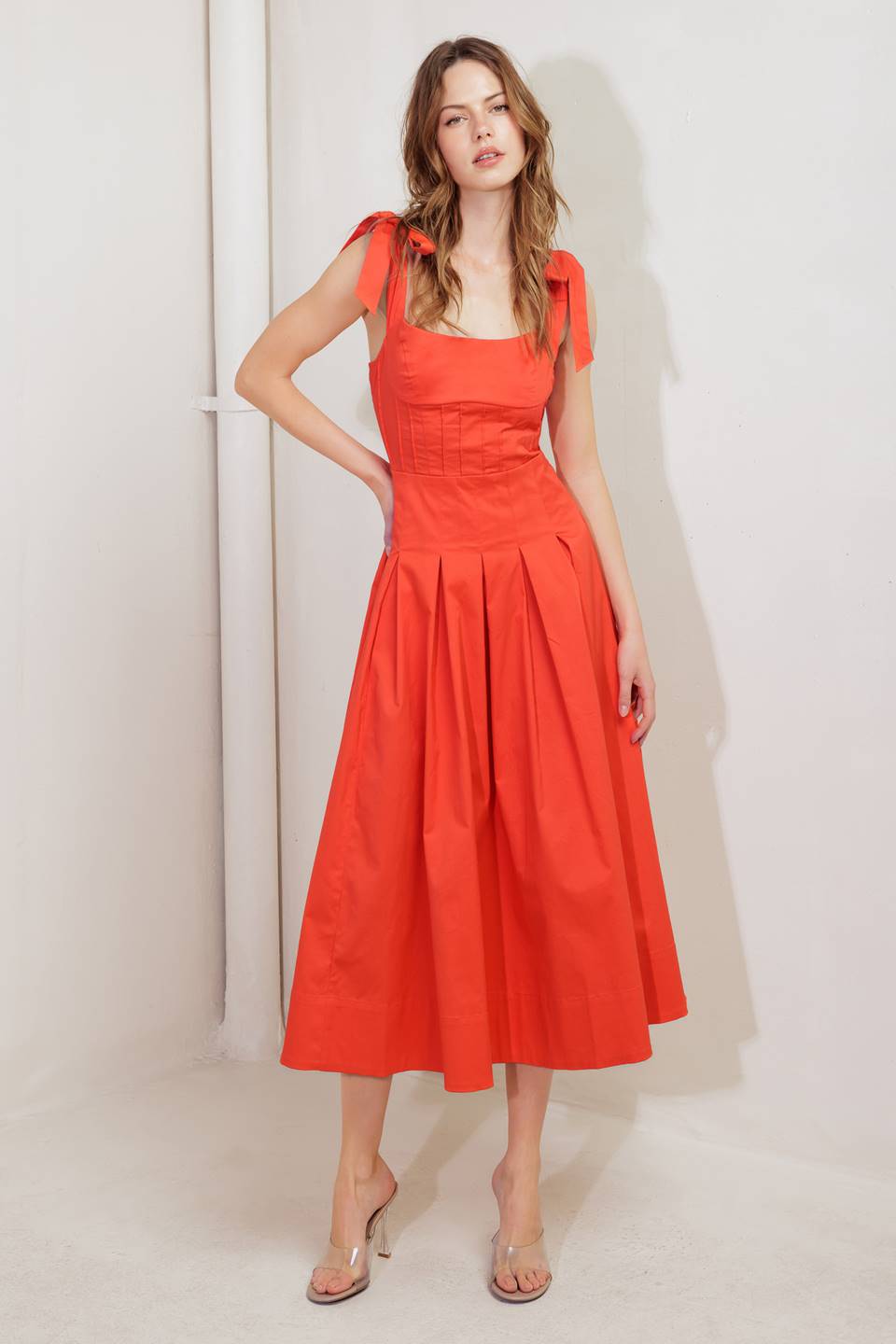A solid red woven midi dress featuring straight neckline, shoulder ties, pintuck waist, inverted pleated skirt and bare back with straps and zipper closure