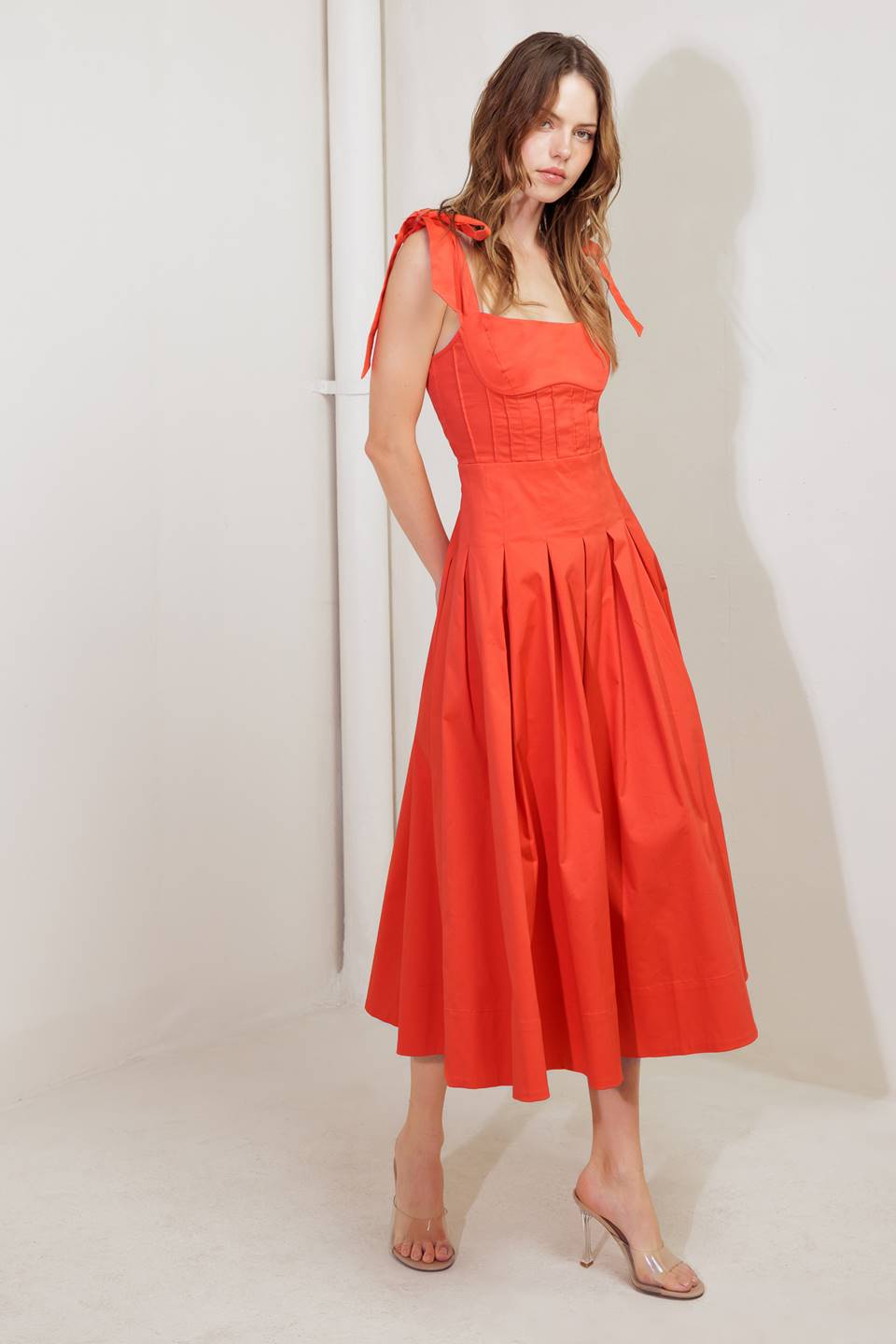 A solid red woven midi dress featuring straight neckline, shoulder ties, pintuck waist, inverted pleated skirt and bare back with straps and zipper closure