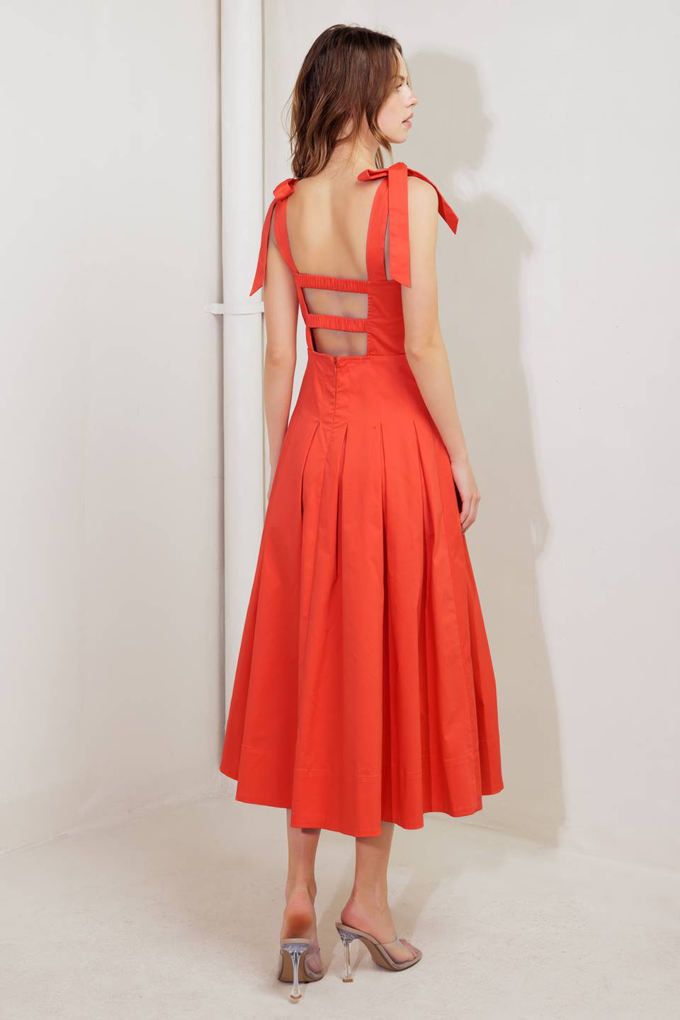 A solid red woven midi dress featuring straight neckline, shoulder ties, pintuck waist, inverted pleated skirt and bare back with straps and zipper closure