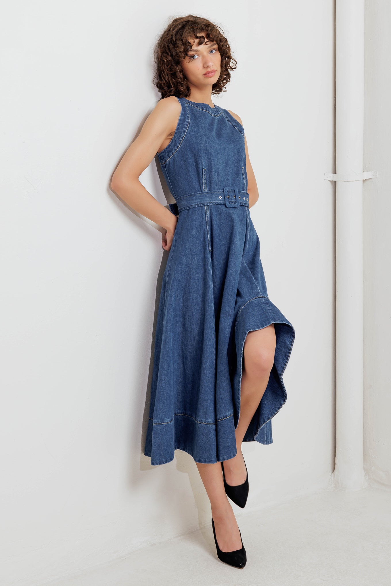 A washed denim midi dress featuring round neckline, sleeveless, self belt, ruffled hemline with side hi-lo and back zipper closure