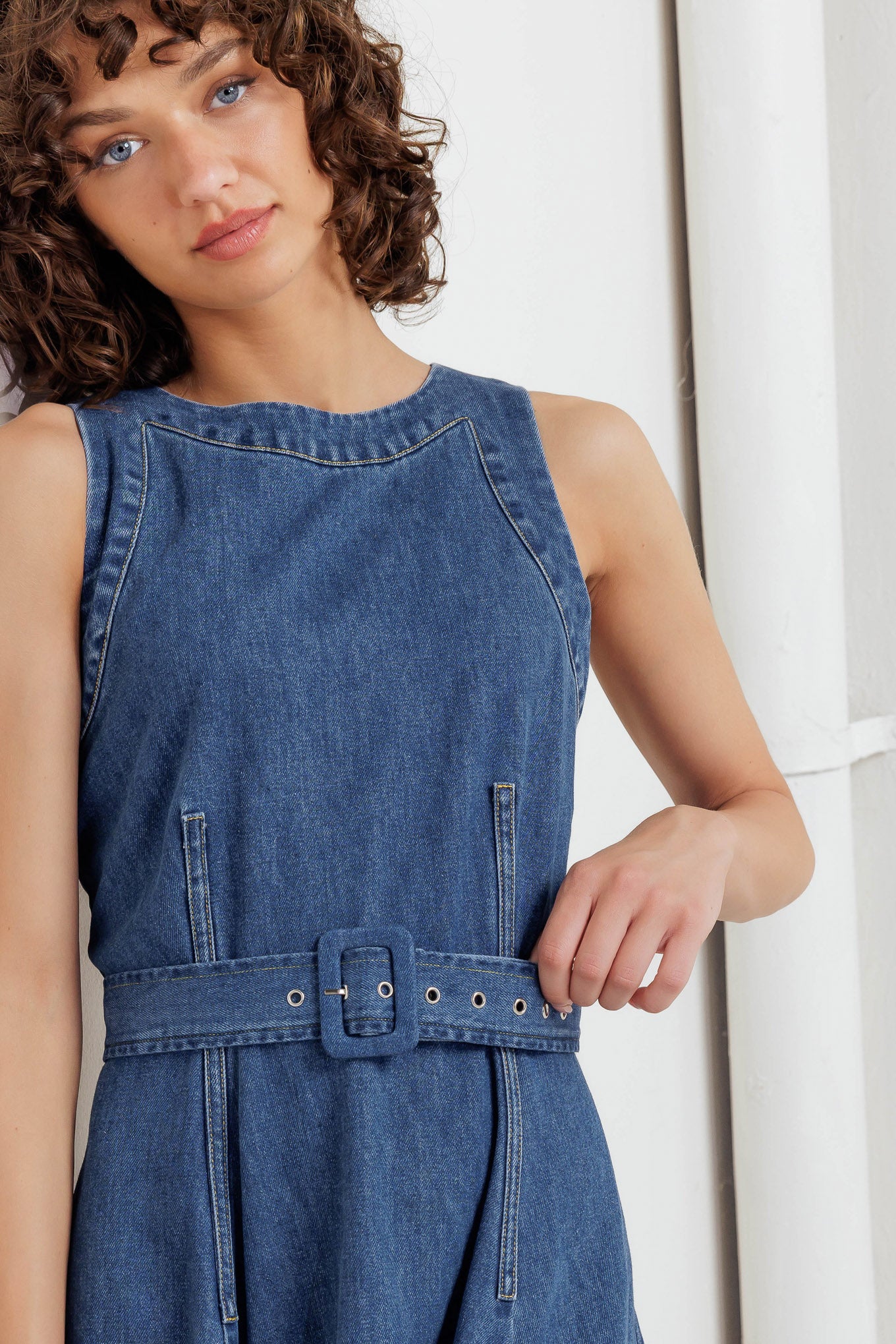 A washed denim midi dress featuring round neckline, sleeveless, self belt, ruffled hemline with side hi-lo and back zipper closure