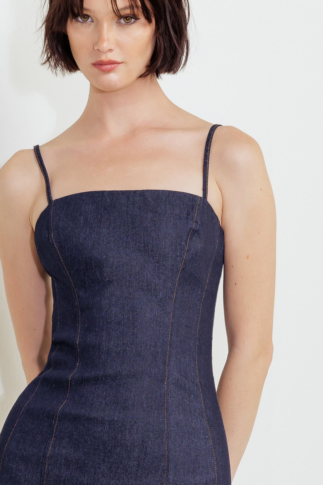 A dark denim midi dress featuring straight neckline, straps, body contouring and back zipper closure with slit