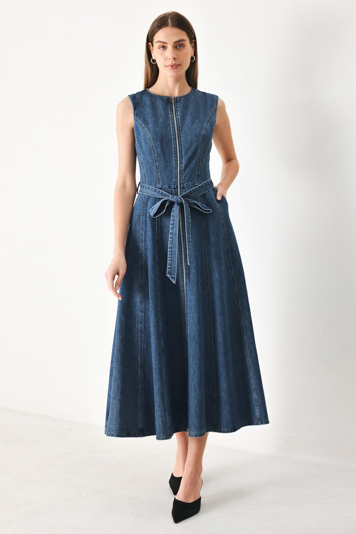Woman wearing a blue denim midi dress featuring round neckline, sleeveless, self sash tie, circle skirt, side pockets and front zipper closure | FREEMARKET | Flying Tomato