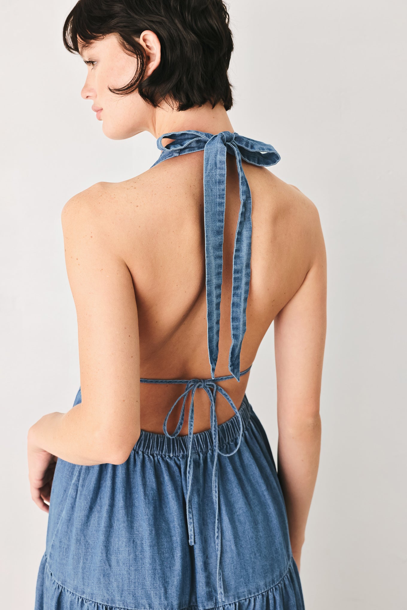 Woman wearing a blue denim midi dress featuring halter neckline, elastic waist, tier skirt, and bare back with tie | FREEMARKET