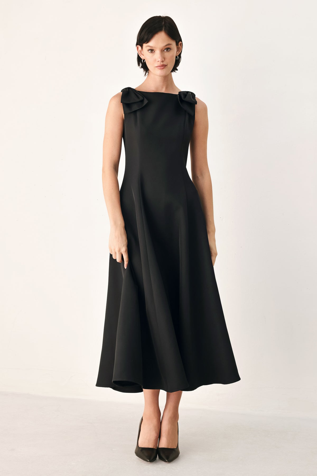 A woman modeling a sleeveless black midi dress with a boat neckline and large bow details on the shoulders | FREEMARKET