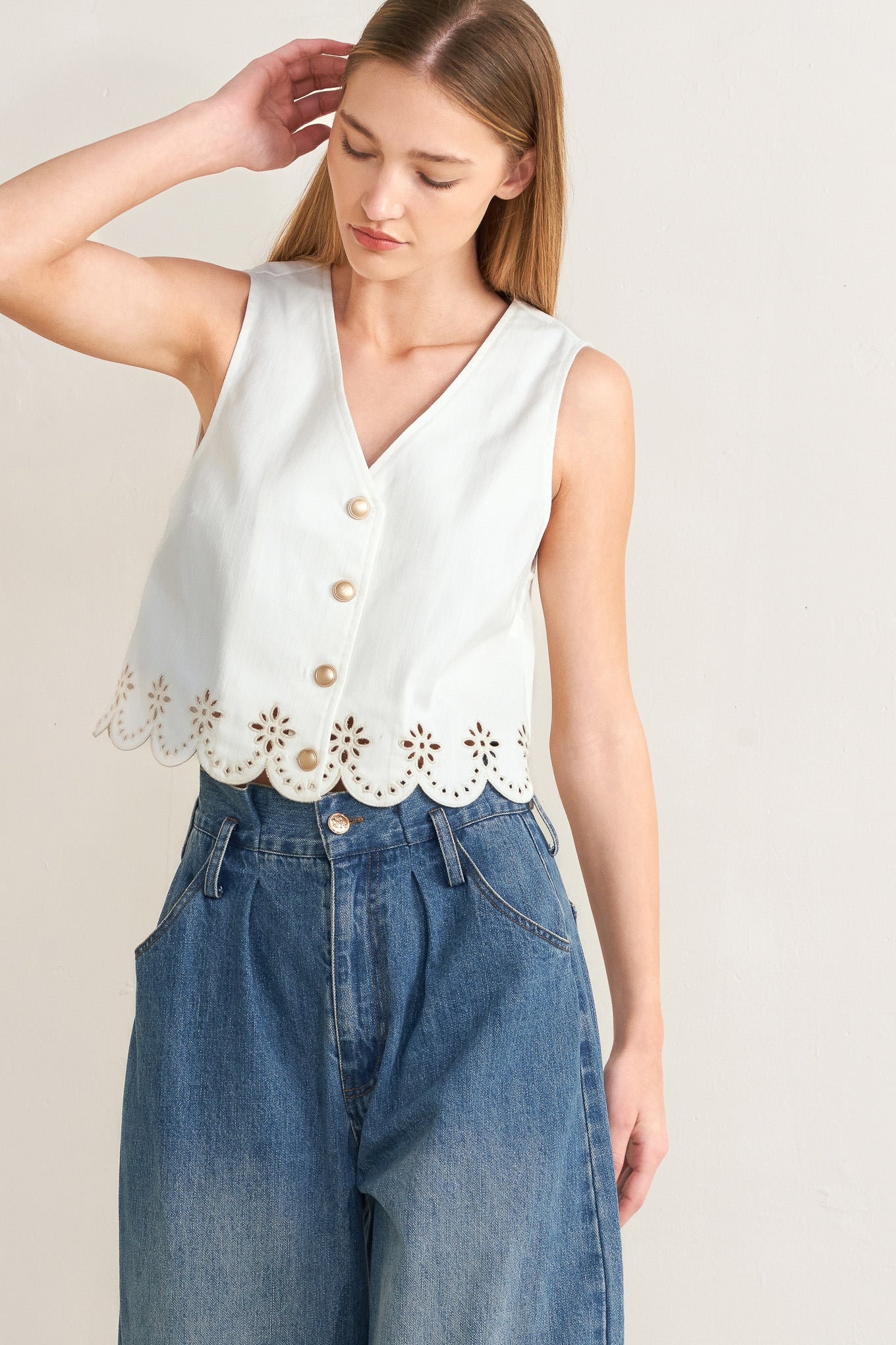 Woman wearing a white sleeveless top with floral cut-out details and blue jeans on a plain background