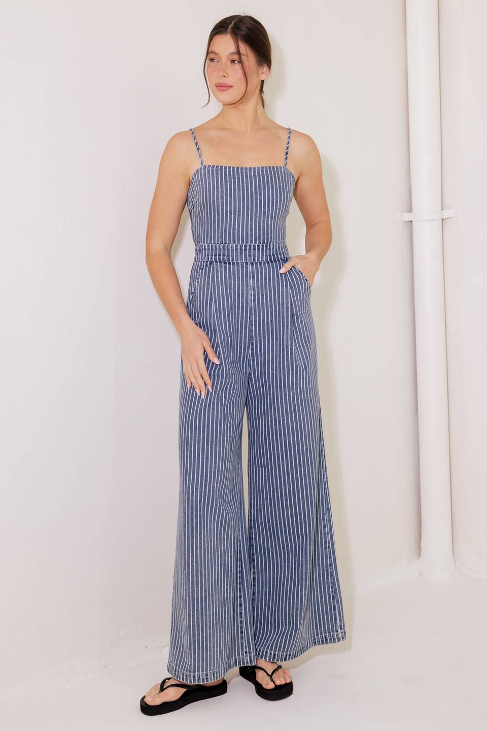 A washed striped denim jumpsuit featuring straight neckline, straps, side pockets, wide leg and bare back with lace up and zipper closure