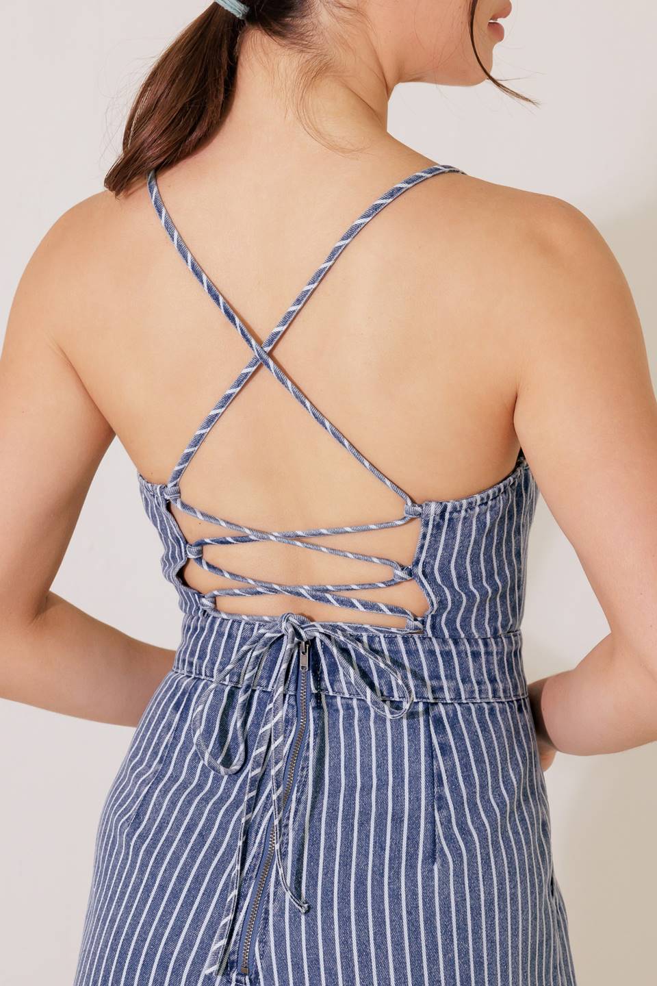 A washed striped denim jumpsuit featuring straight neckline, straps, side pockets, wide leg and bare back with lace up and zipper closure