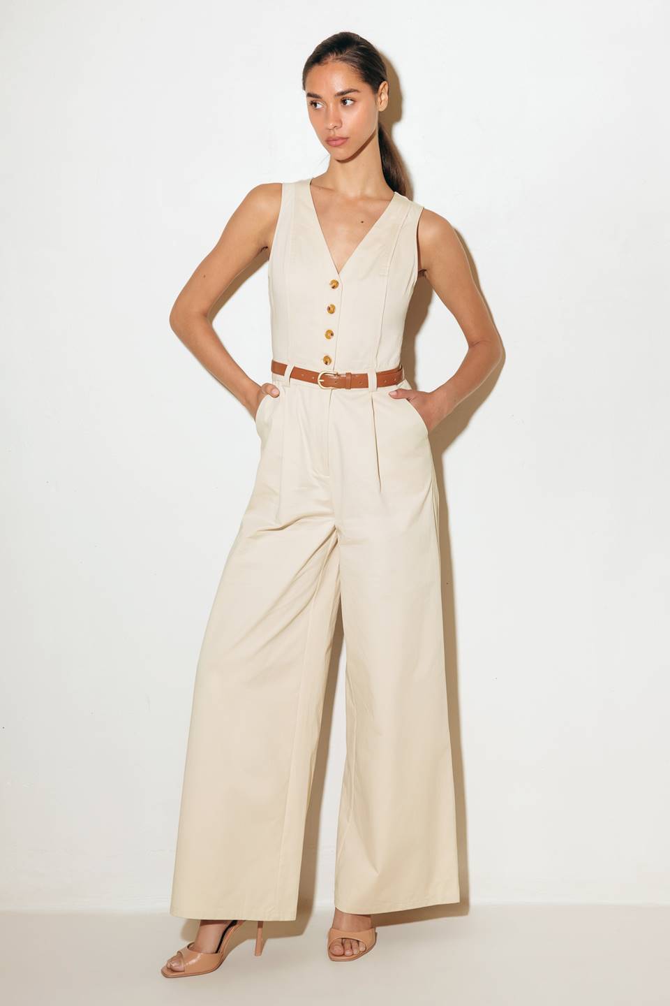 A solid woven jumpsuit featuring V neckline, sleeveless, front button/zipper closure, belt, side pockets, wide leg and surplice back with cut out