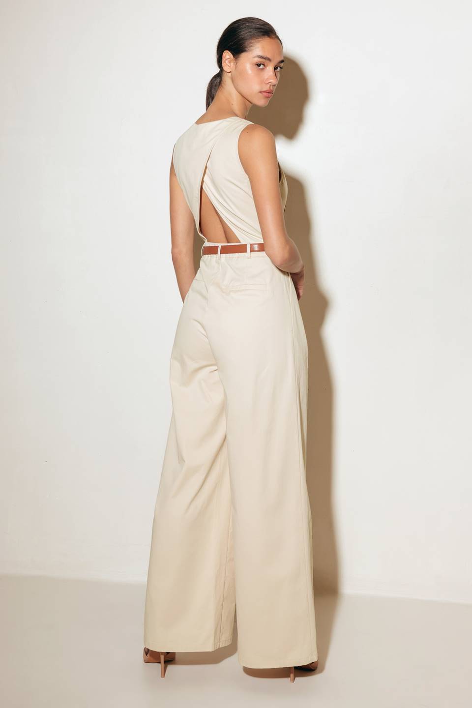 A solid woven jumpsuit featuring V neckline, sleeveless, front button/zipper closure, belt, side pockets, wide leg and surplice back with cut out