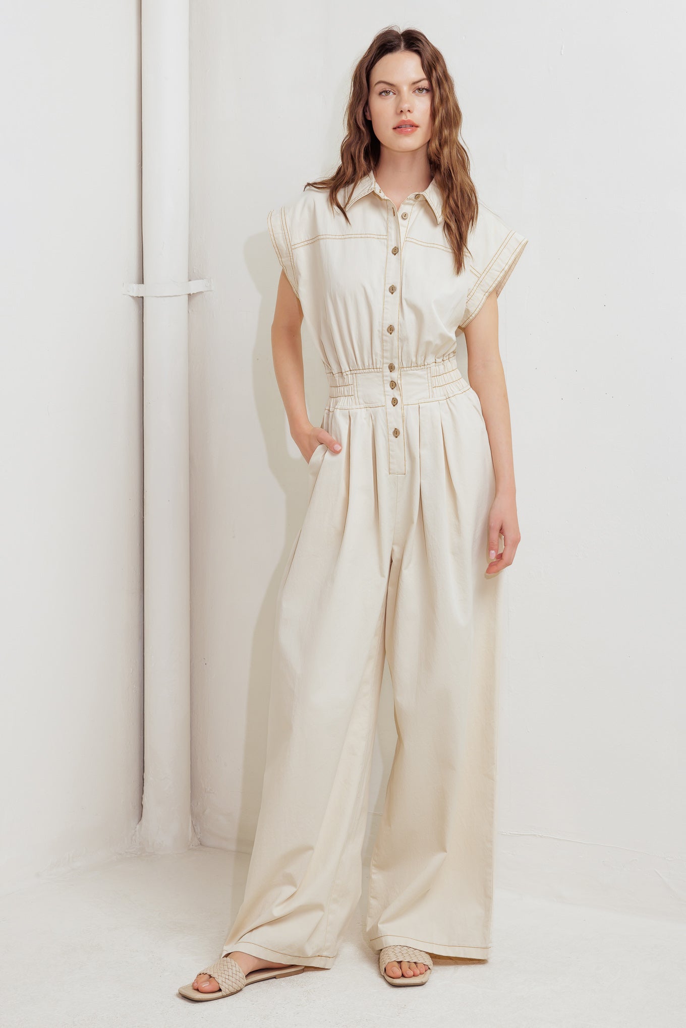 A solid woven jumpsuit featuring collar, dropped shoulder with short sleeve, button down, elasticized waistband, side pockets and wide leg