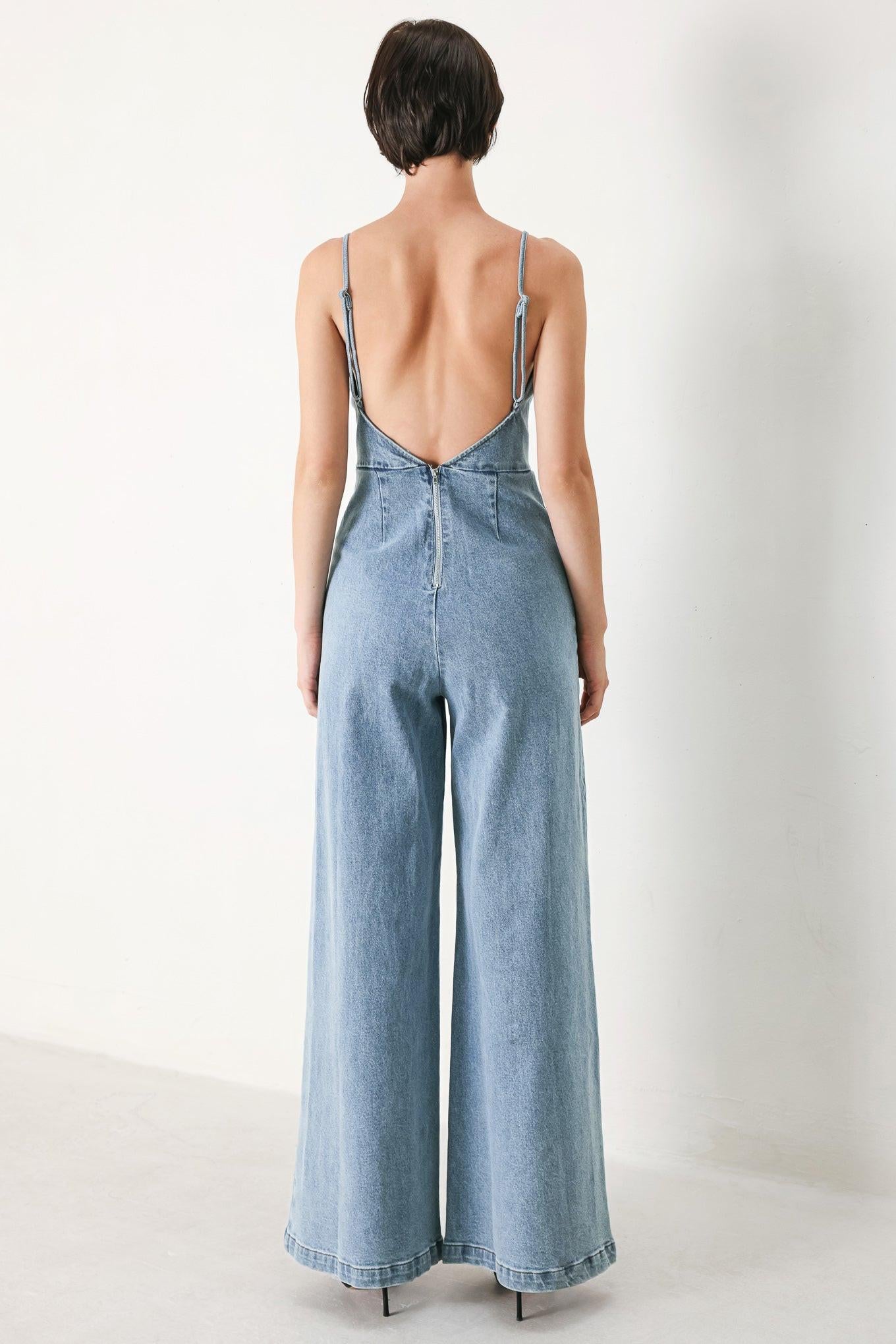 A washed denim jumpsuit featuring U neckline, straps, waist yoke, wide leg, bare back with zipper closure