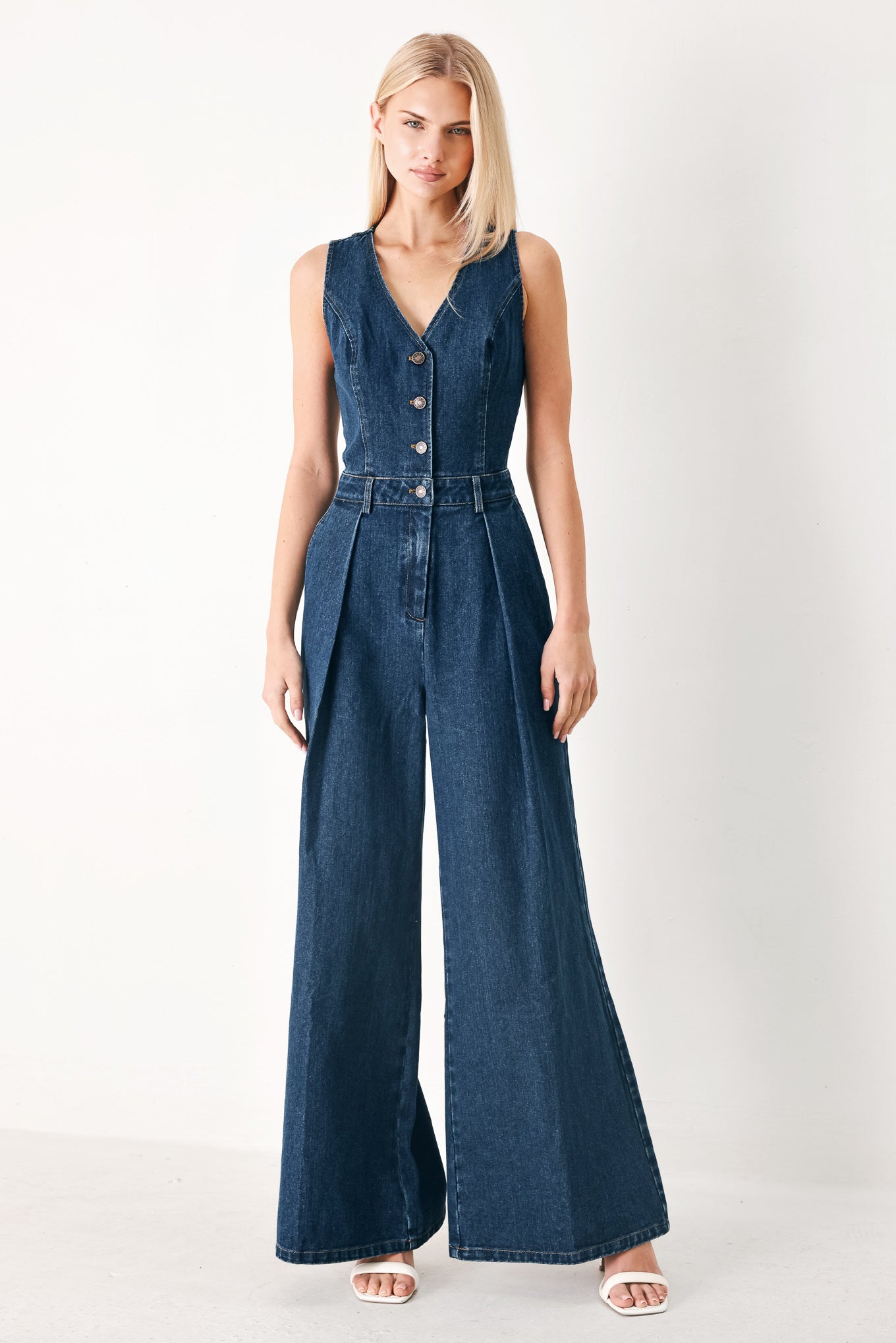 Woman wearing a denim jumpsuit featuring V neckline, sleeveless, front button closure, side pockets, wide leg and surplice back with cut out | FREEMARKET