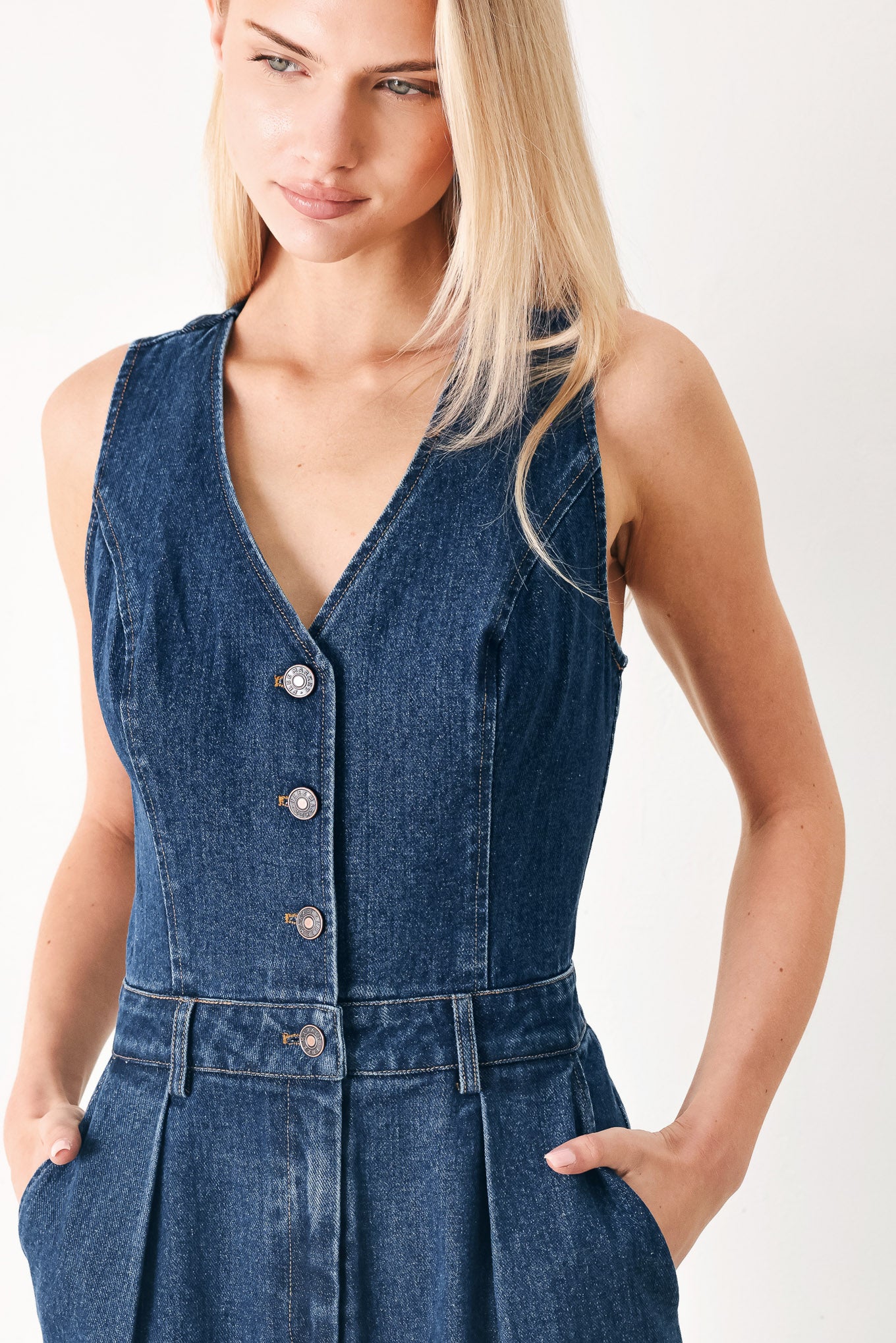Woman wearing a denim jumpsuit featuring V neckline, sleeveless, front button closure, side pockets, wide leg and surplice back with cut out | FREEMARKET