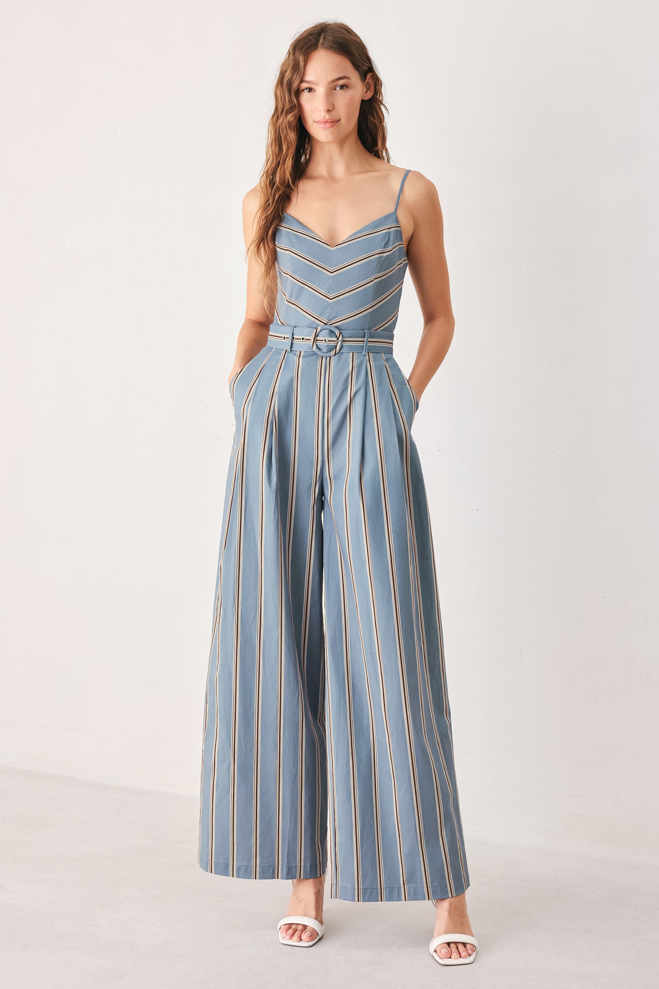 A woman wearing a sleeveless wide-leg jumpsuit with vertical blue, beige, and white stripes, featuring a V-neck bodice, thin shoulder straps, and a matching fabric belt with a silver buckle | FREEMARKET
