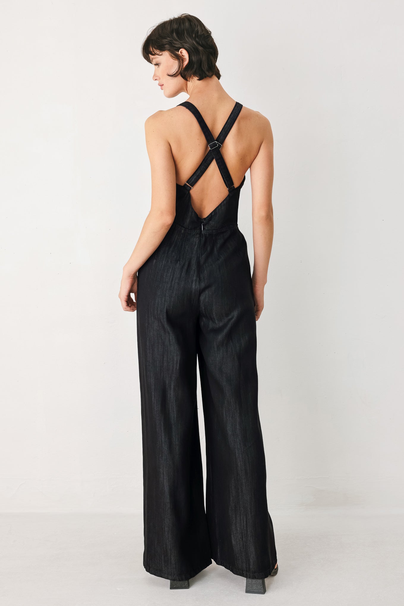 Woman wearing a black denim jumpsuit featuring halter neckline with plunging V and hardware, pockets, wide leg, bare back with cross straps and zipper closure | FREEMARKET