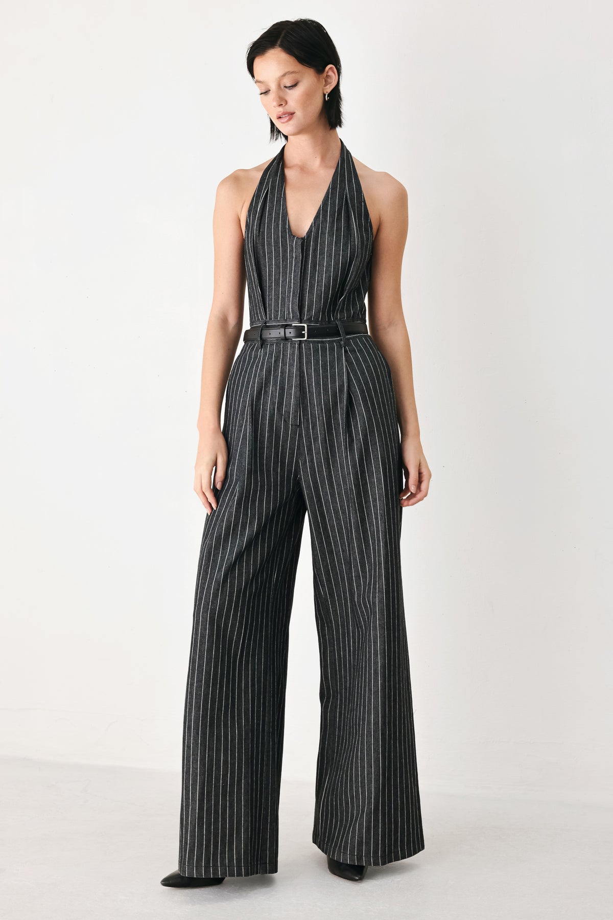 A model wearing a sleeveless halter-neck jumpsuit with black and white pinstripes, a matching black belt, and wide-leg trousers, standing against a plain white studio background.