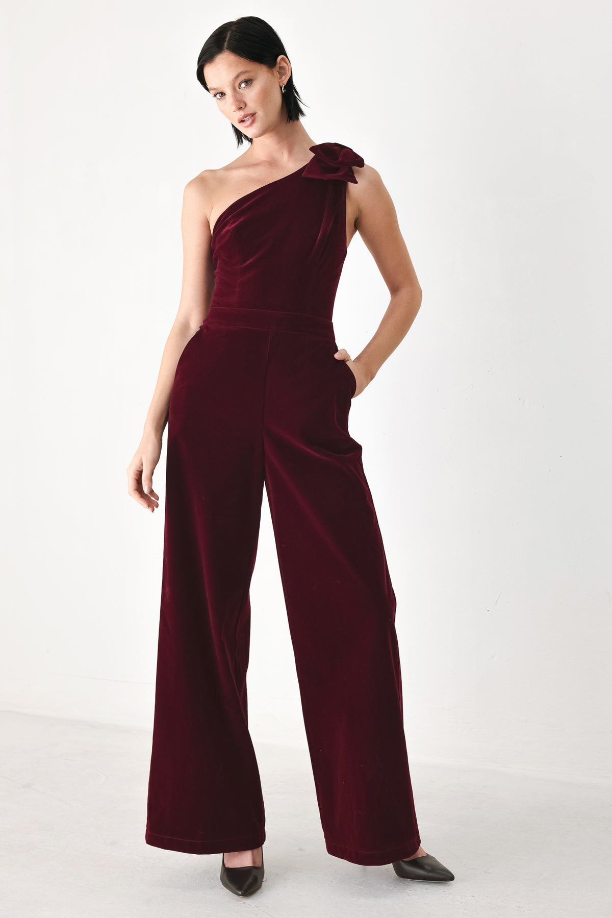 A woman wearing a deep burgundy velvet one-shoulder jumpsuit with a large bow detail on the shoulder | FREEMARKET