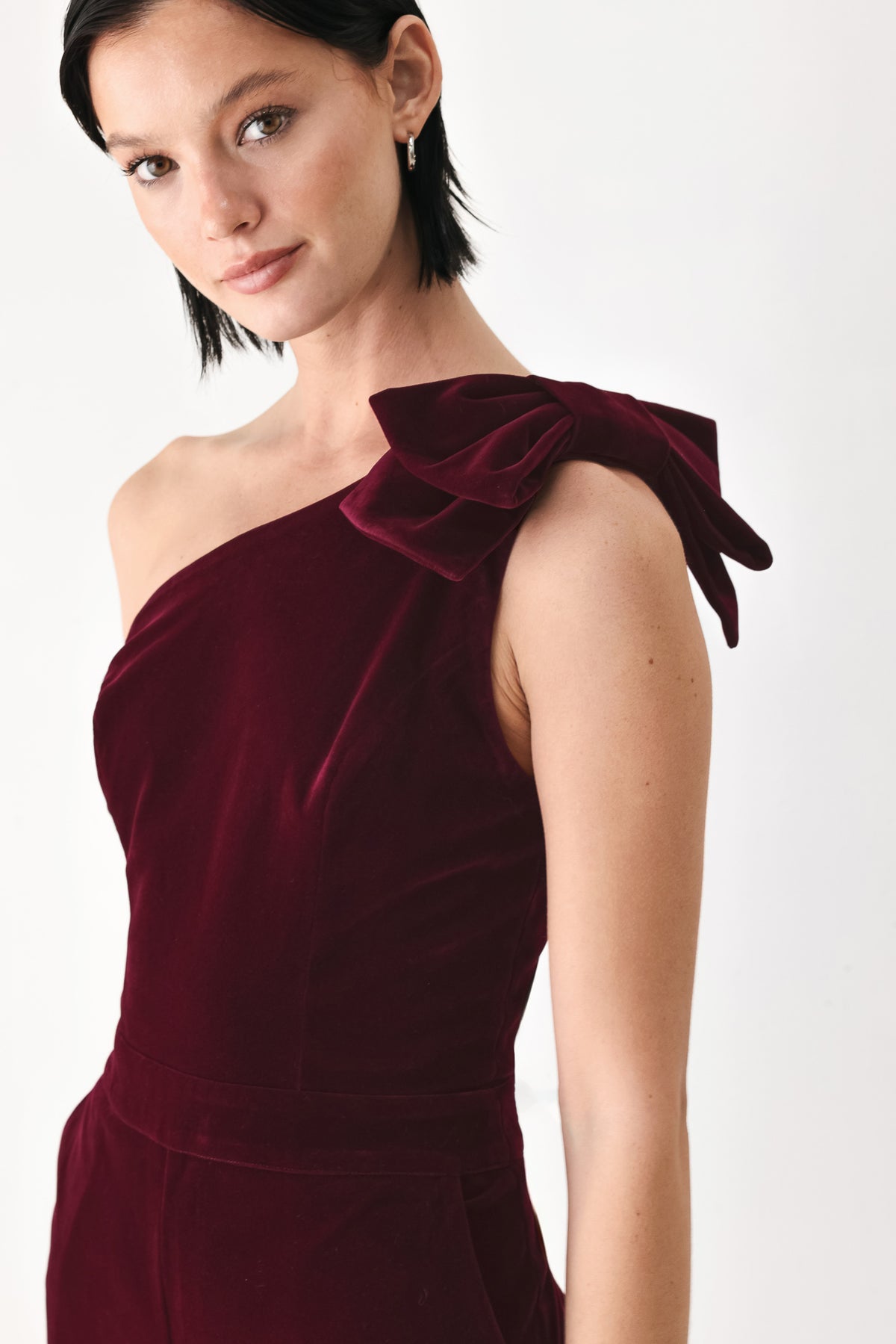 A woman wearing a deep burgundy velvet one-shoulder jumpsuit with a large bow detail on the shoulder | FREEMARKET