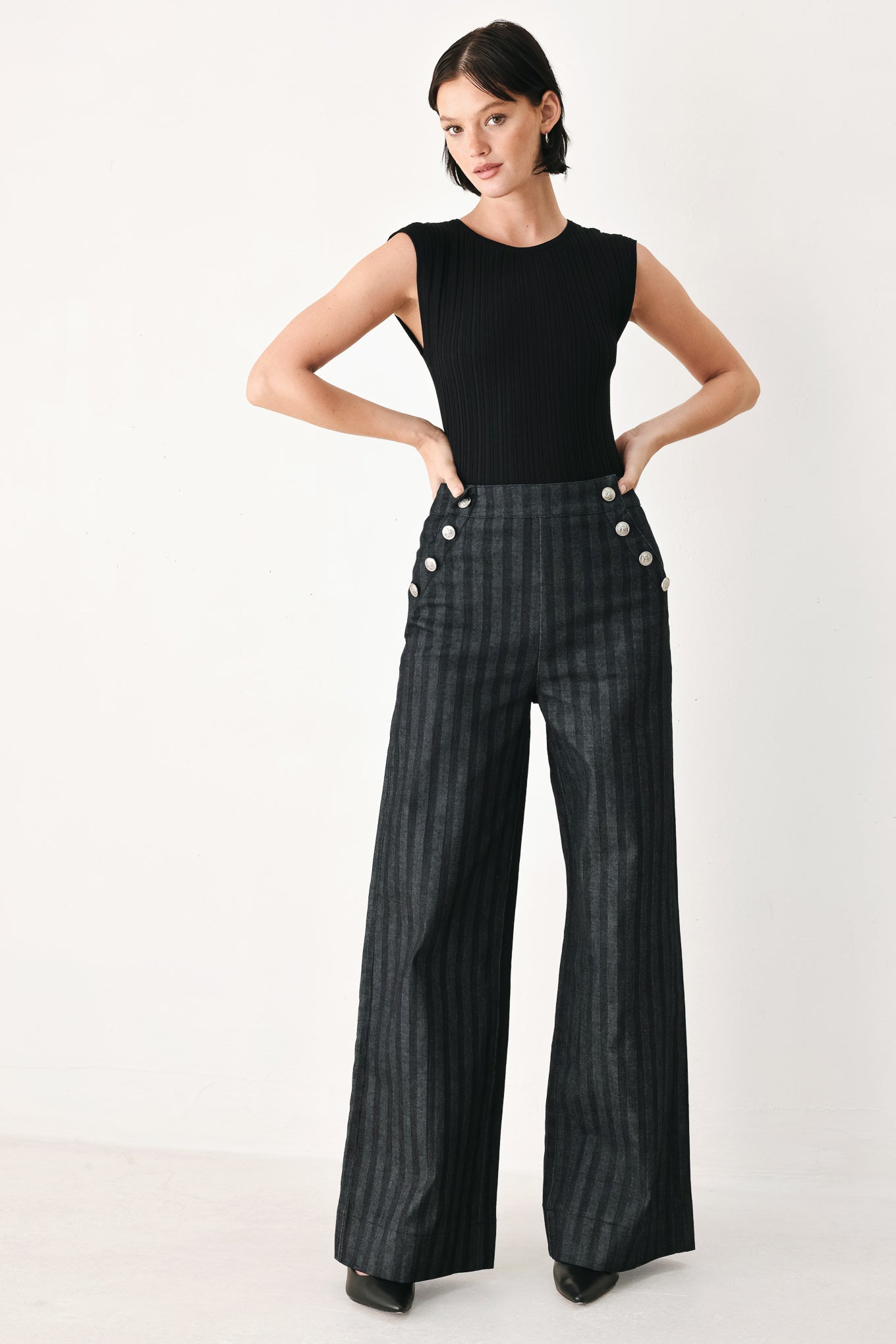 A model wearing a pair of high-waisted, wide-leg trousers in a dark gray and black pinstripe pattern, featuring decorative silver buttons on the front waistband | FREEMARKET