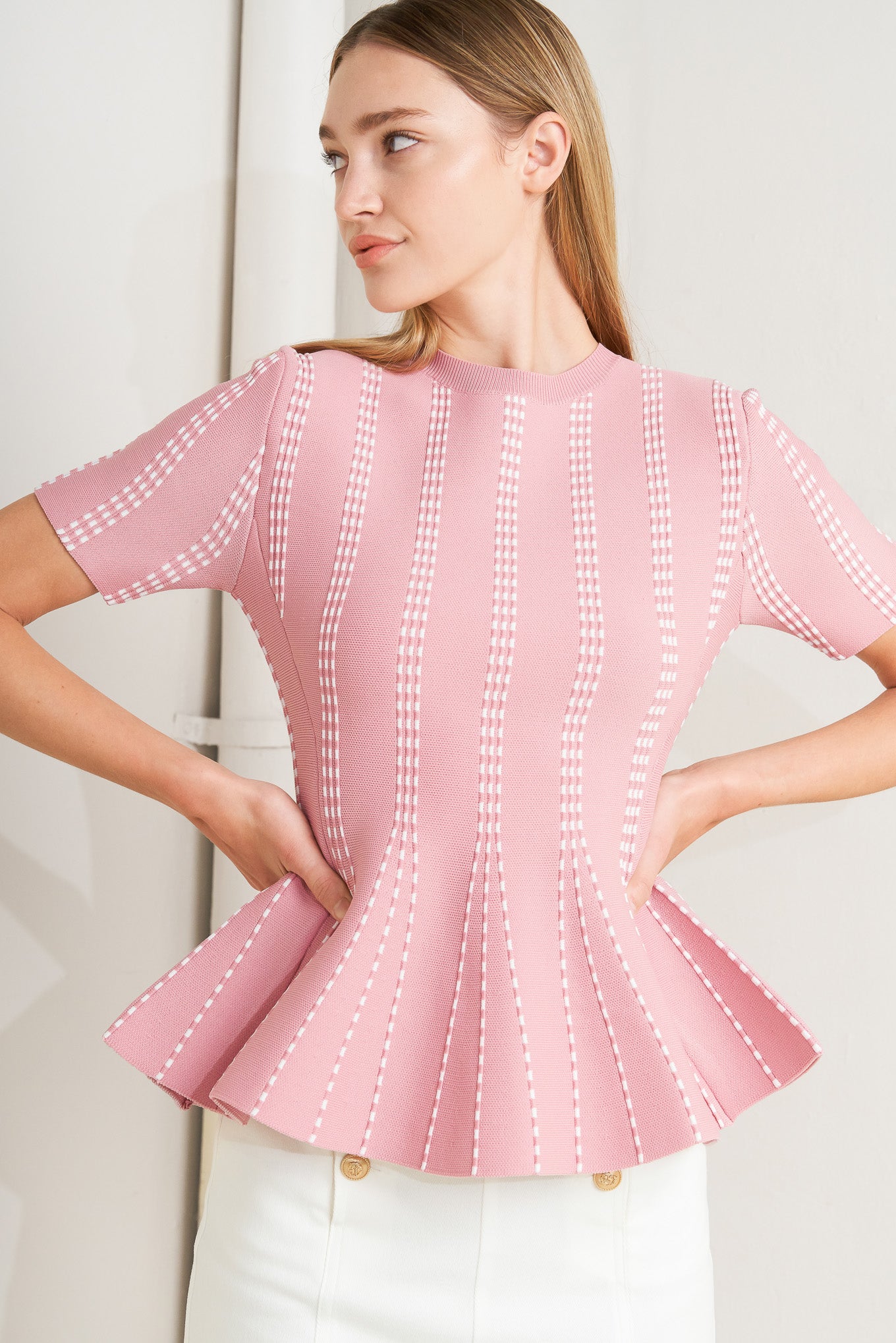 Woman wearing a pink blouse with white stitching against a neutral background AND A jacquard sweater top featuring round neckline, short sleeve and peplum FLYING TOMATO