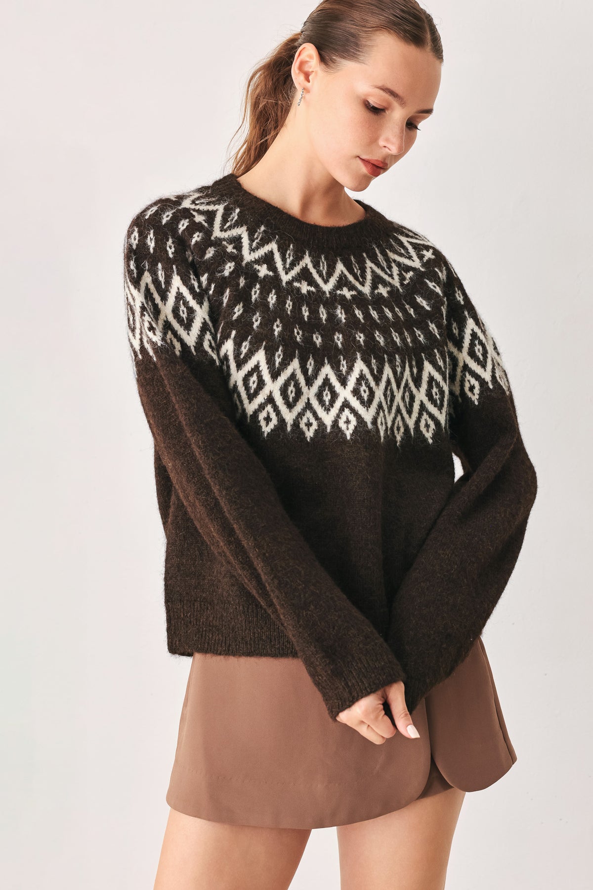 A model stands against a light gray studio background wearing a dark brown knit sweater with a white geometric pattern across the chest | FREEMARKET