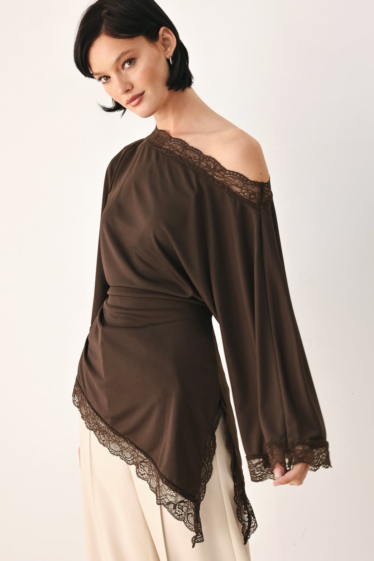 A model wearing a dark brown asymmetrical off-the-shoulder top with brown lace trim along the neckline, cuffs, and hem | FREEMARKET