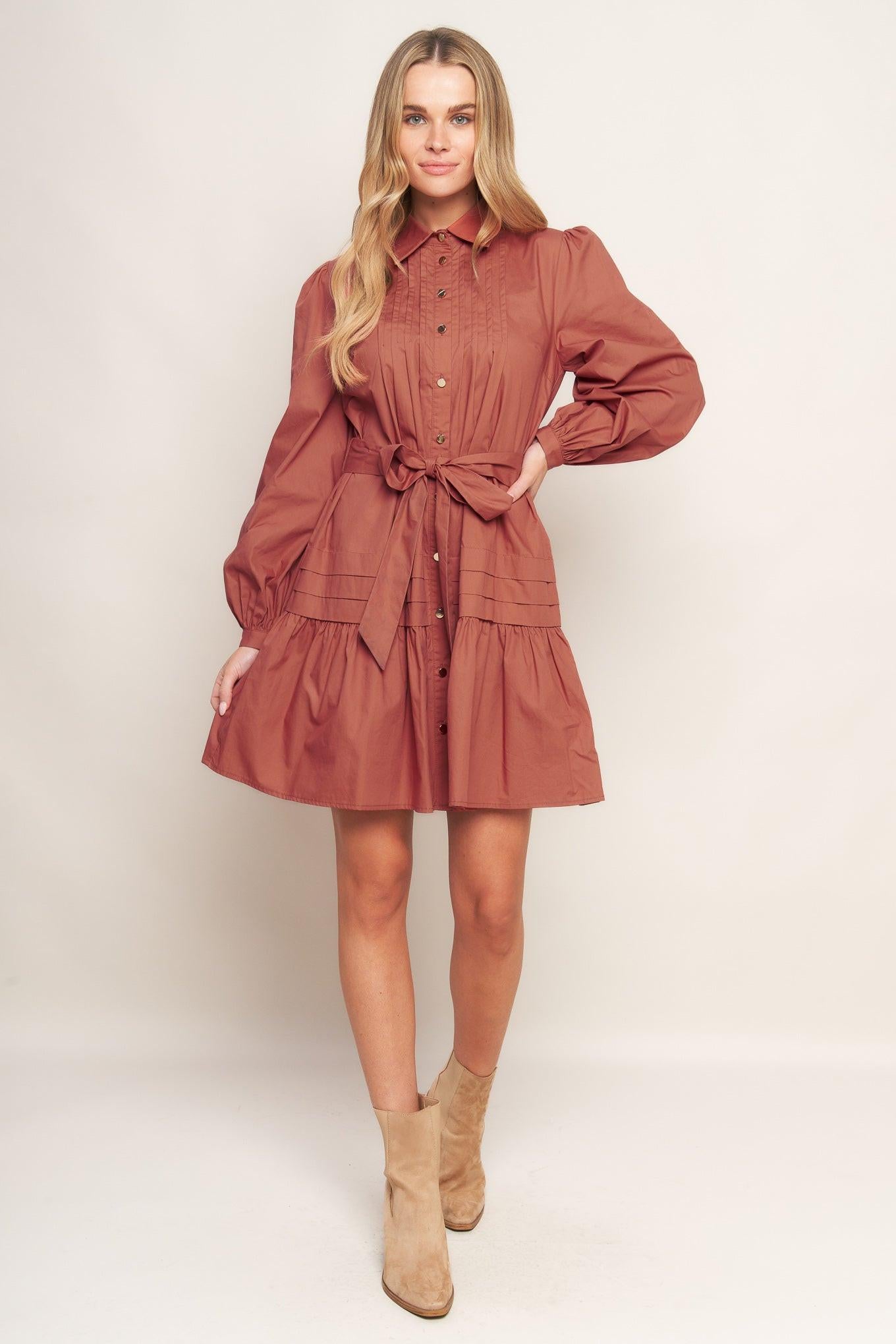 A solid woven mini dress featuring a shirt collar, button down, front pleats, long sleeve, self-sash tie, and ruffled hemline