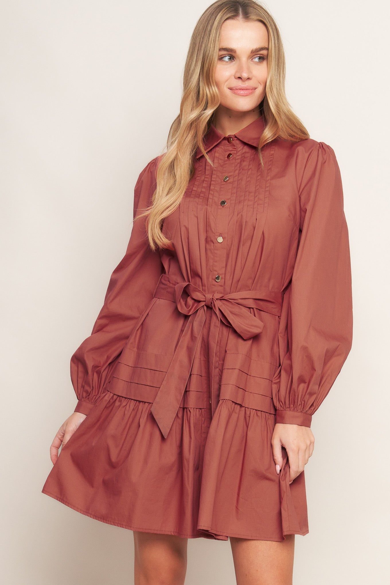 A solid woven mini dress featuring a shirt collar, button down, front pleats, long sleeve, self-sash tie, and ruffled hemline