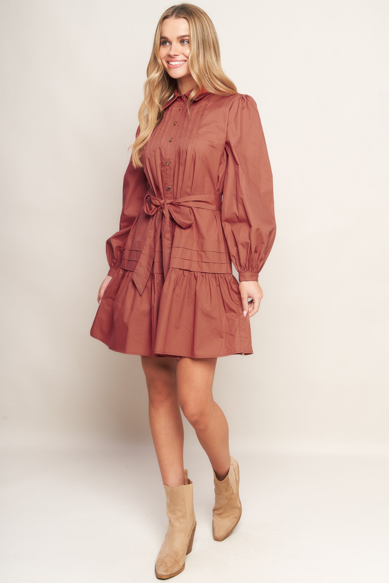 A solid woven mini dress featuring a shirt collar, button down, front pleats, long sleeve, self-sash tie, and ruffled hemline