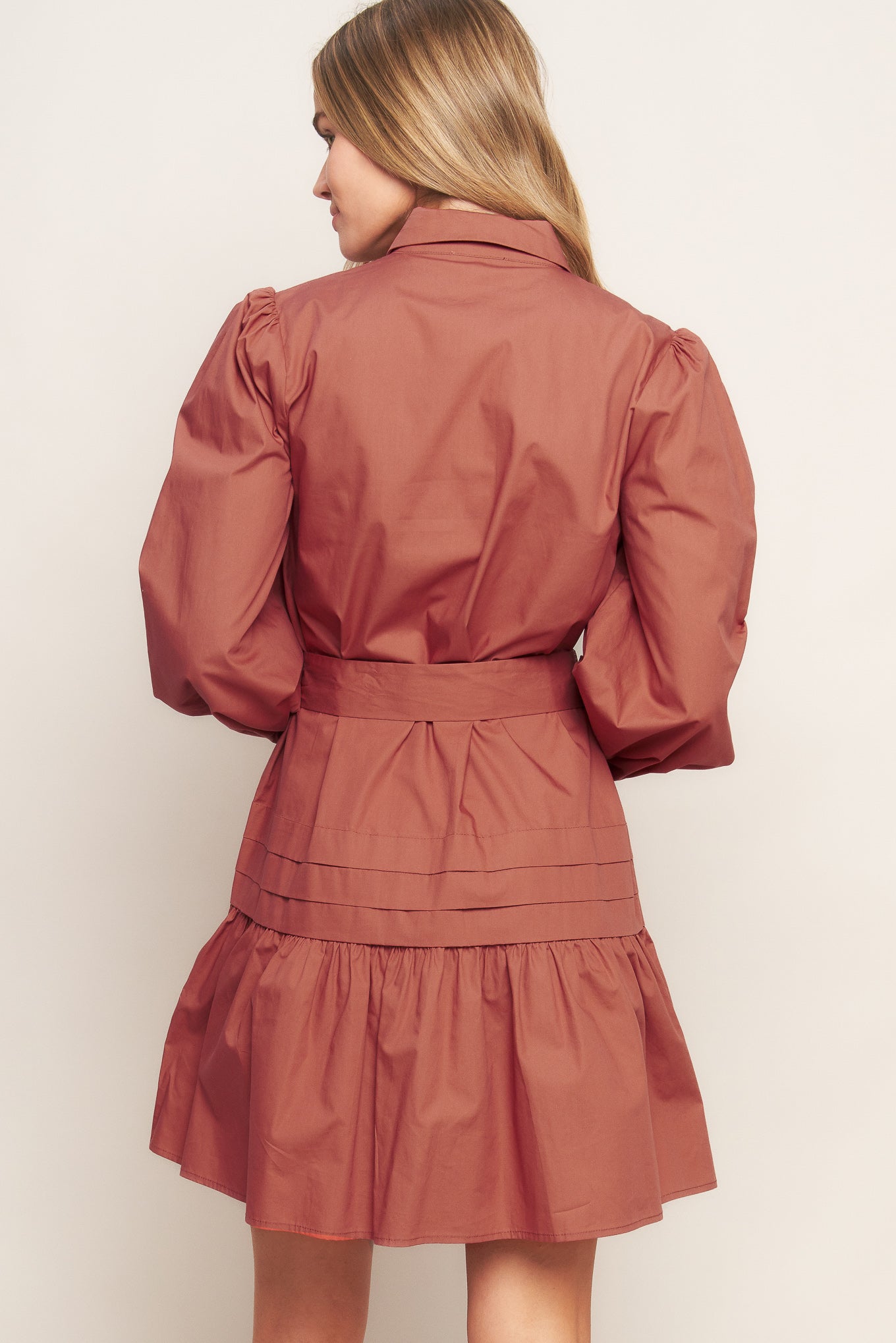 A solid woven mini dress featuring a shirt collar, button down, front pleats, long sleeve, self-sash tie, and ruffled hemline