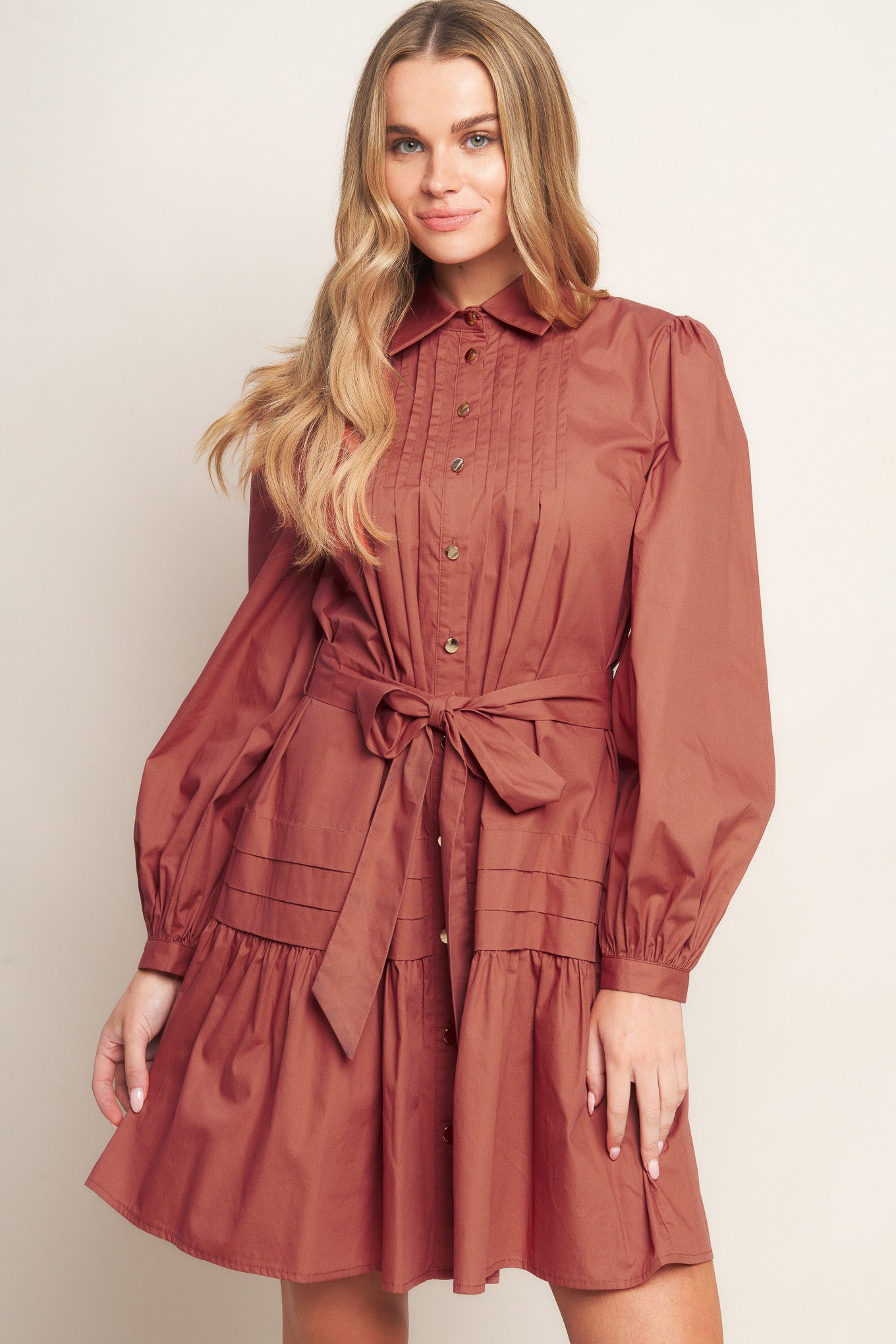 A solid woven mini dress featuring a shirt collar, button down, front pleats, long sleeve, self-sash tie, and ruffled hemline