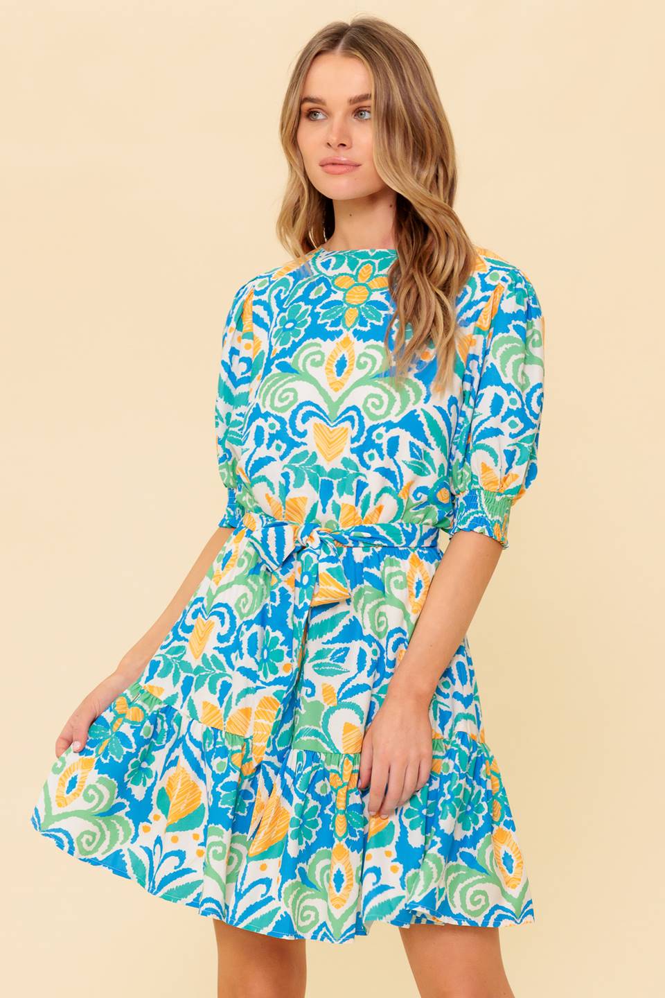 A printed woven mini dress featuring round neckline, short puff sleeve with smocked cuff, elasticized waist with self sash tie, ruffled hemline and back neck button closure