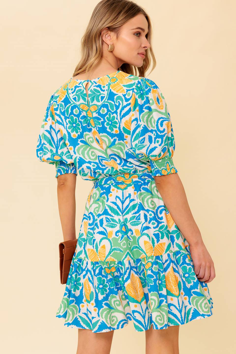 A printed woven mini dress featuring round neckline, short puff sleeve with smocked cuff, elasticized waist with self sash tie, ruffled hemline and back neck button closure