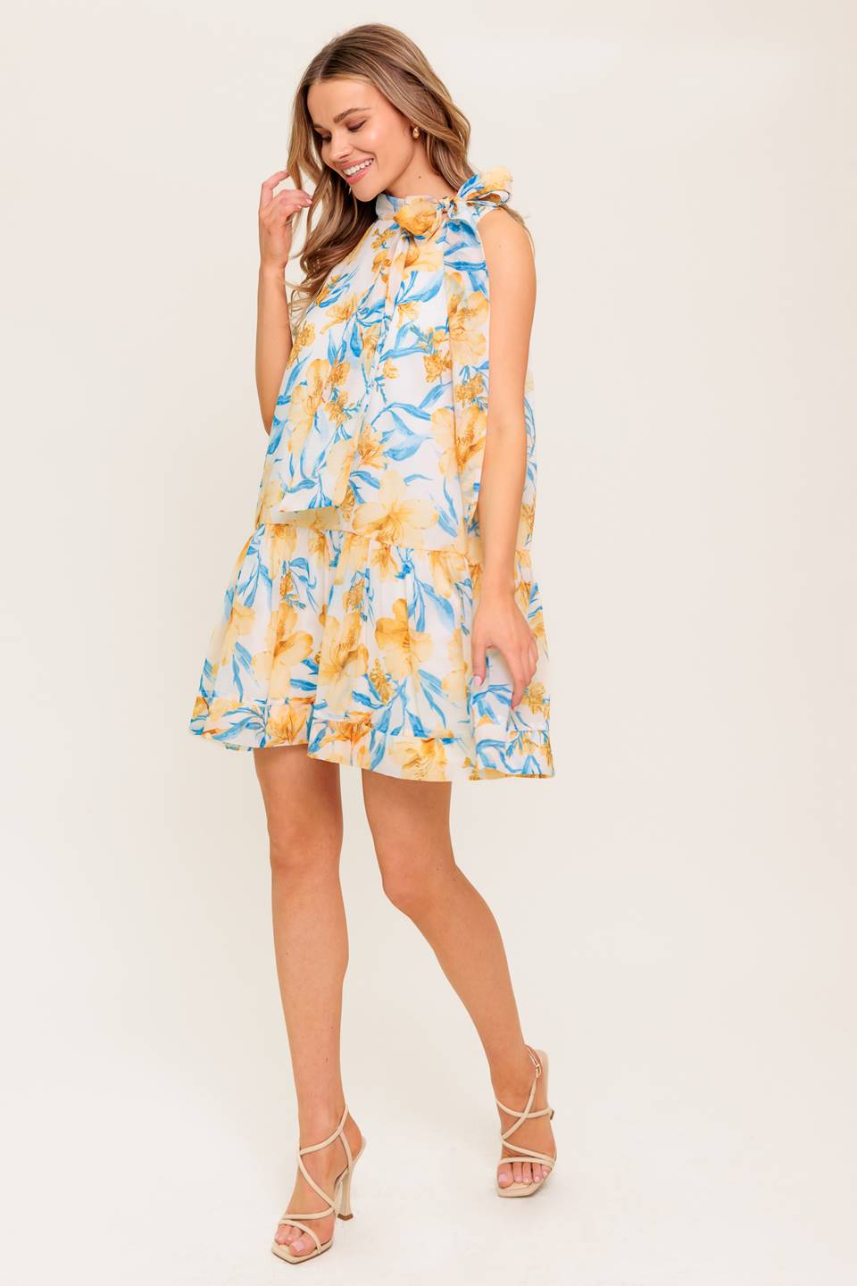 A printed woven mini dress featuring high neckline with bow tie, sleeveless, drop waist, ruffle hemline and back button closure.