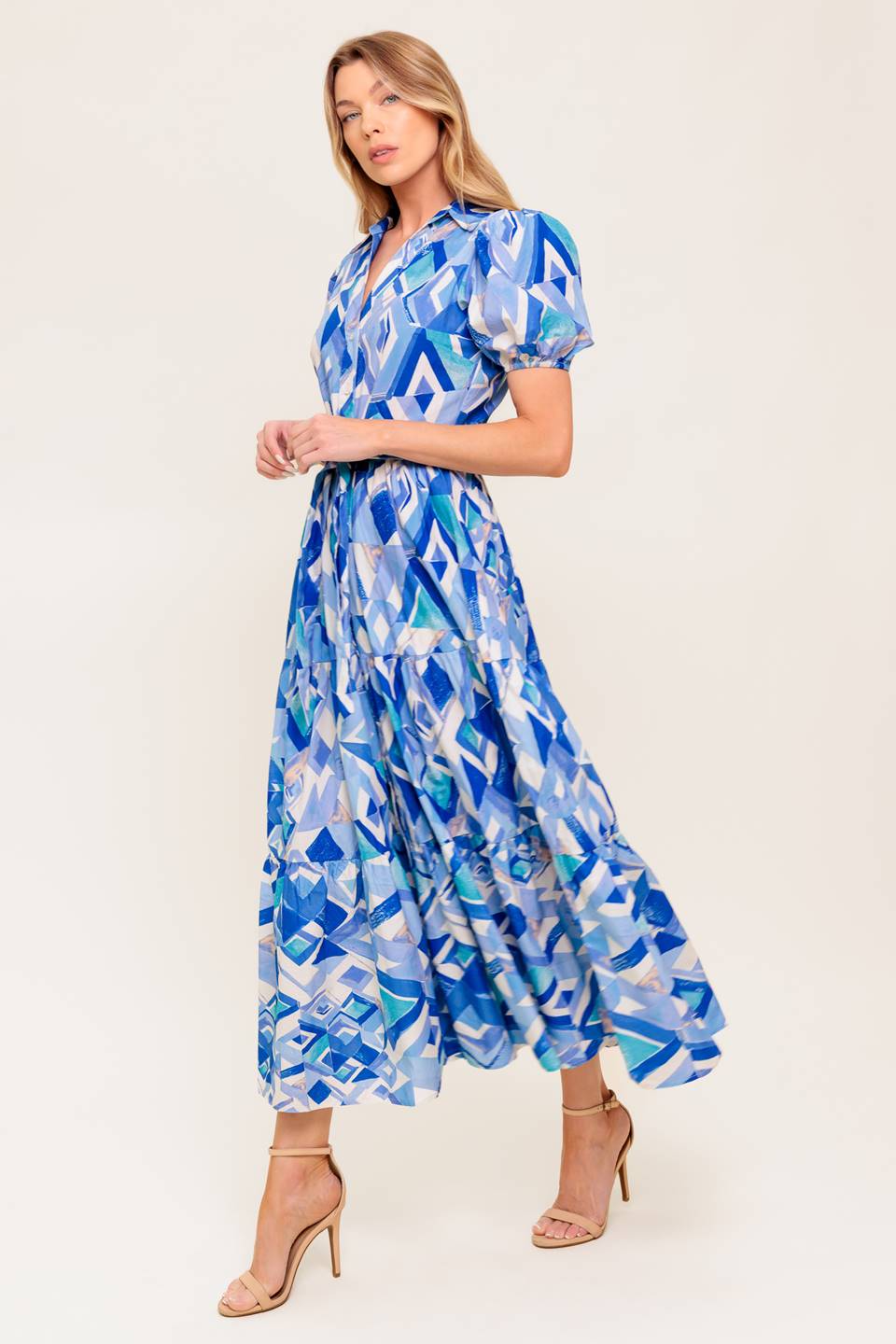 A printed woven midi dress featuring collar, V neck opening, short puff sleeve, front button down closure, self sash tie and tiered skirt.