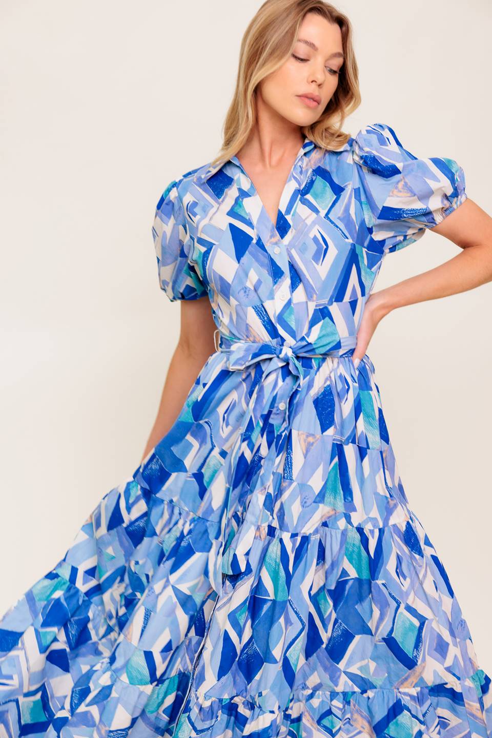A printed woven midi dress featuring collar, V neck opening, short puff sleeve, front button down closure, self sash tie and tiered skirt.