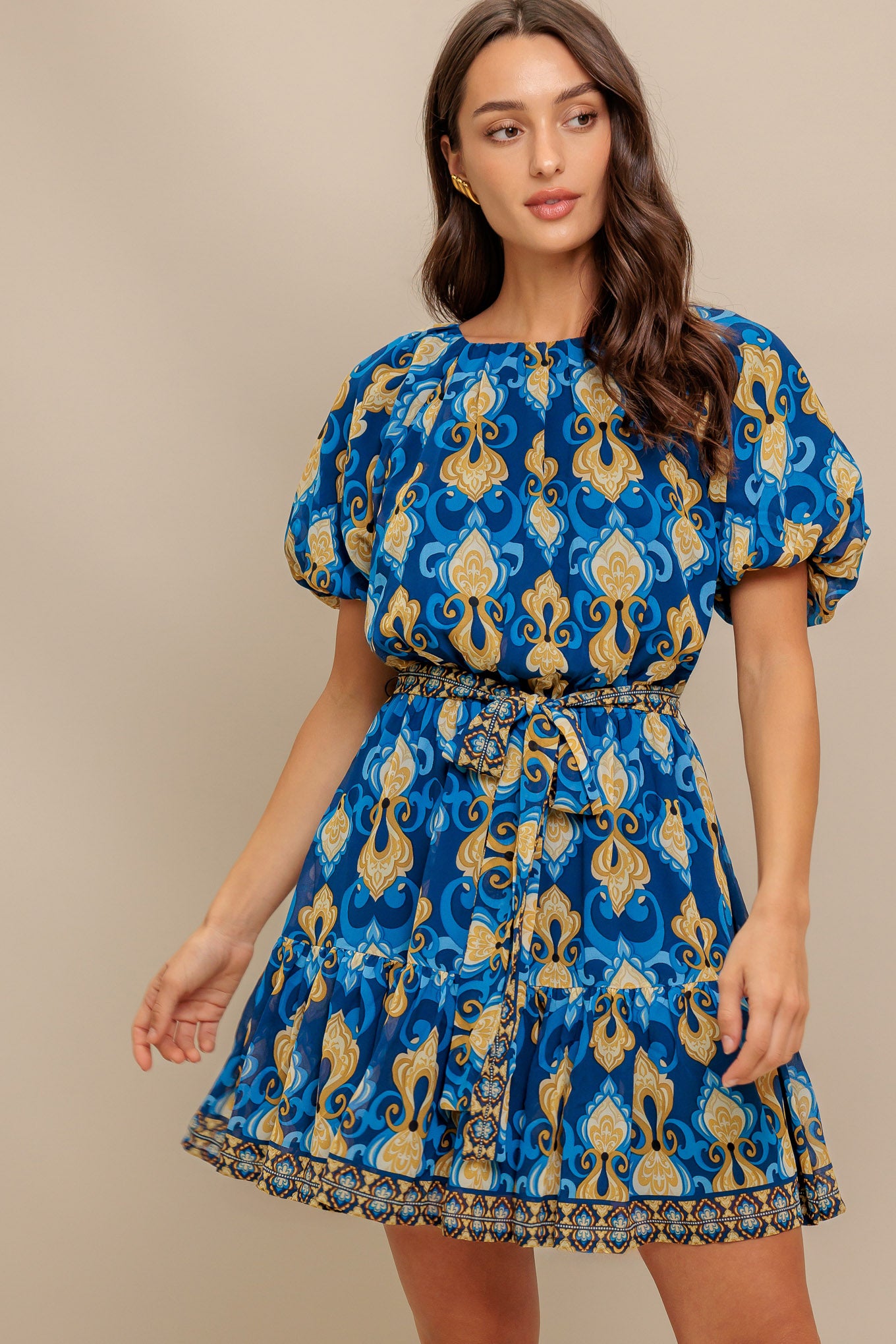 A printed woven mini dress featuring round neckline, short puff sleeve, elasticized waist with self sash, ruffled hemline and back button closure