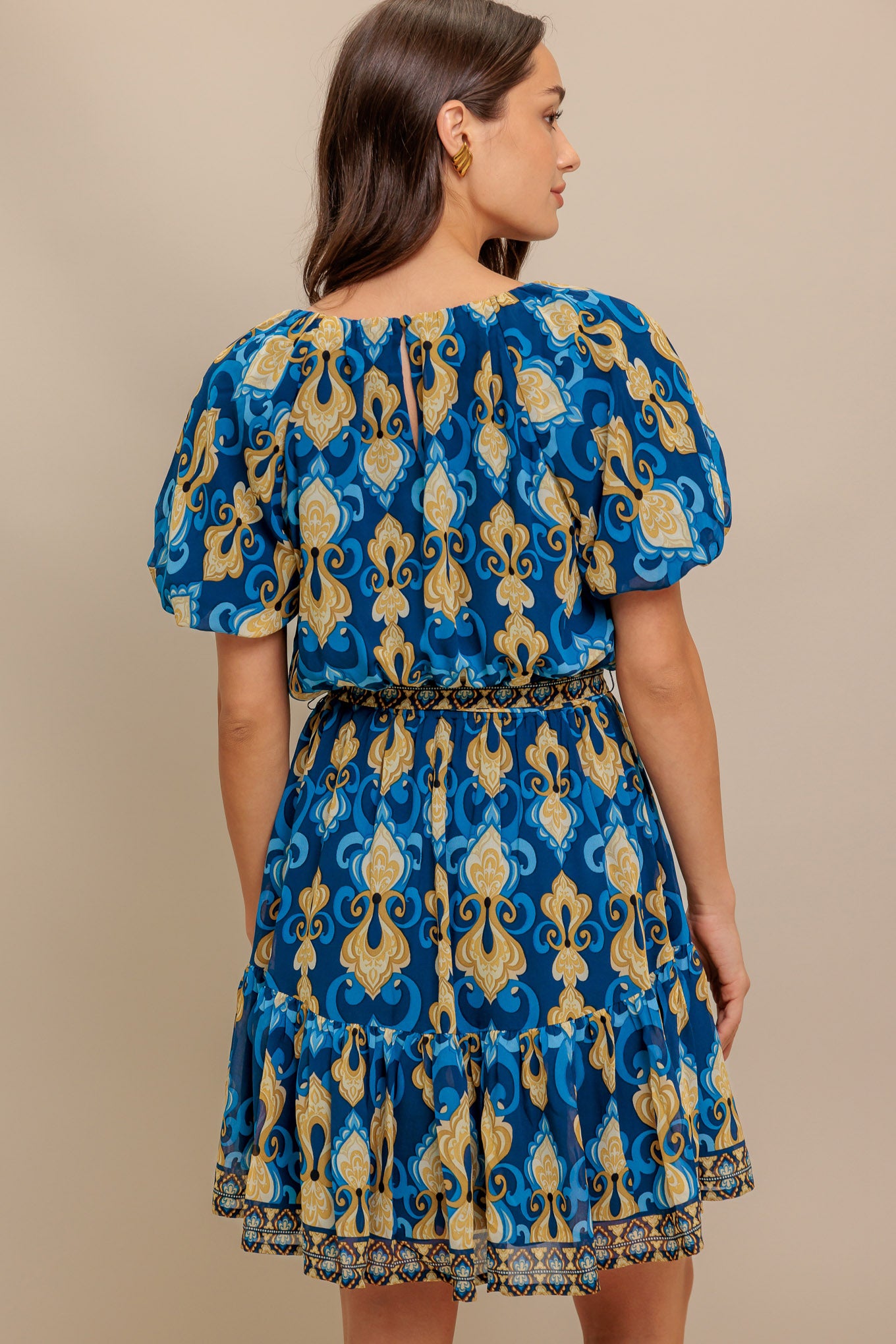 A printed woven mini dress featuring round neckline, short puff sleeve, elasticized waist with self sash, ruffled hemline and back button closure