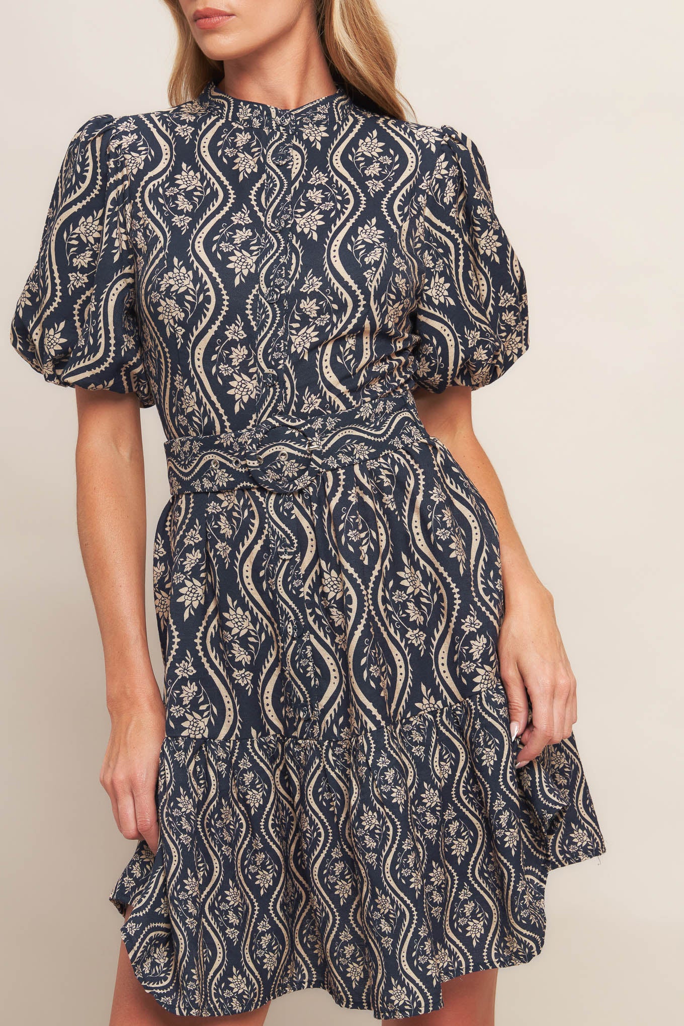 A printed woven mini dress featuring high neckline, short puff sleeve, front button down closure, self belt and ruffled hemline