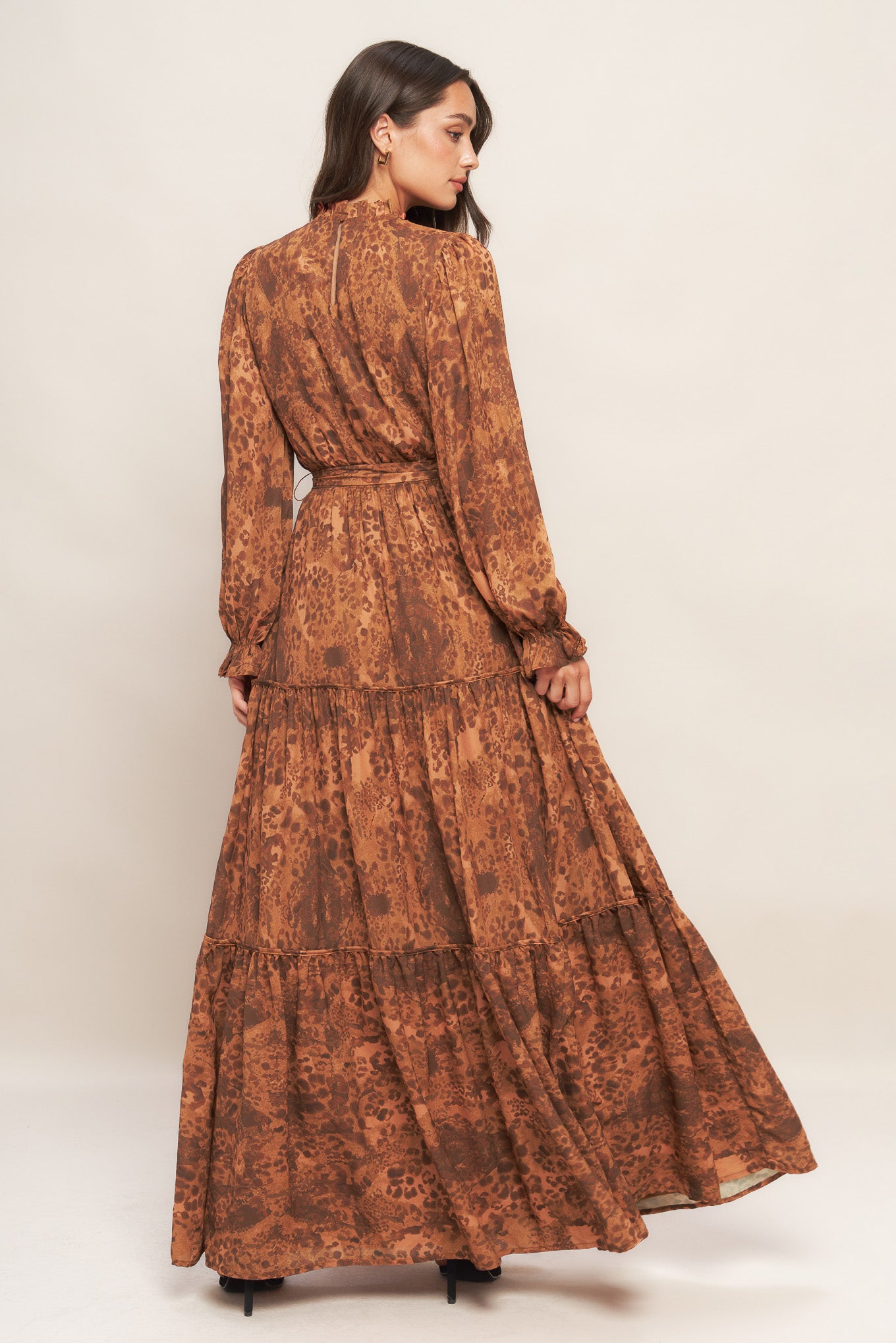 A printed woven maxi dress featuring ruffled high neckline, long sleeve, elasticized waist, self sash tie, tiered skirt and back neck button closure