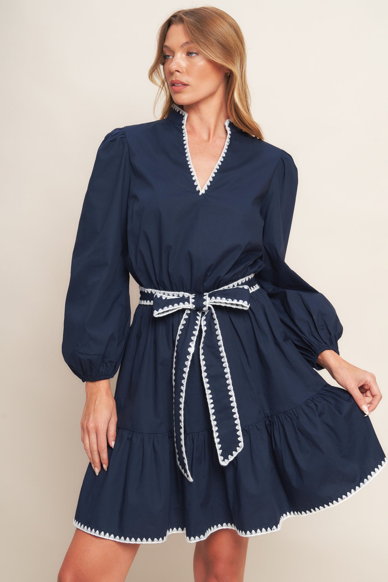 A solid woven mini dress featuring V neckline, long sleeve, elasticized waist, self sash tie, ruffled hemline and embroidered edge detail