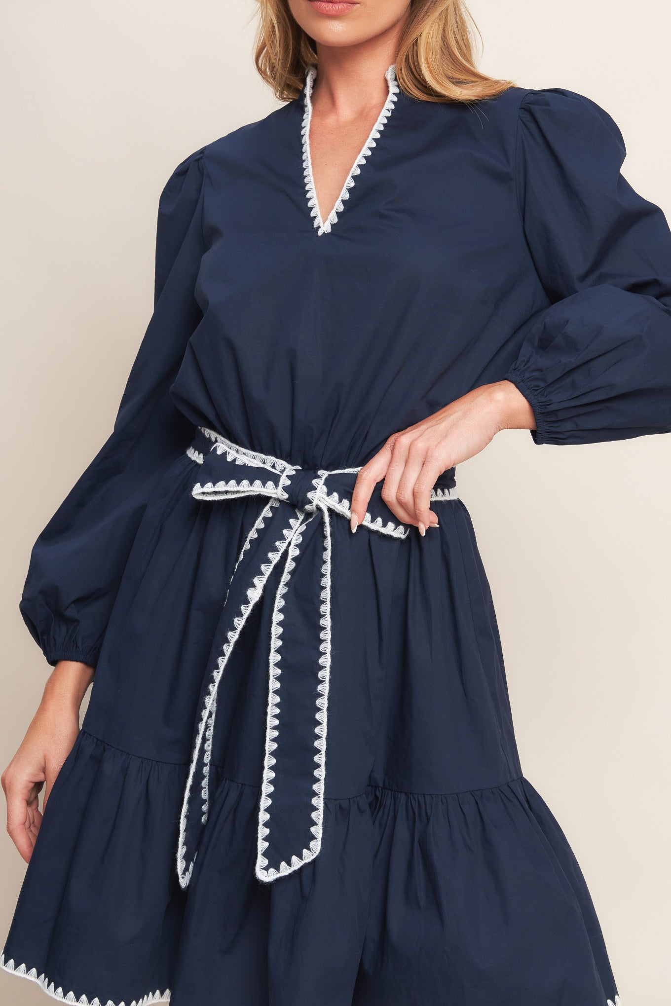 A solid woven mini dress featuring V neckline, long sleeve, elasticized waist, self sash tie, ruffled hemline and embroidered edge detail