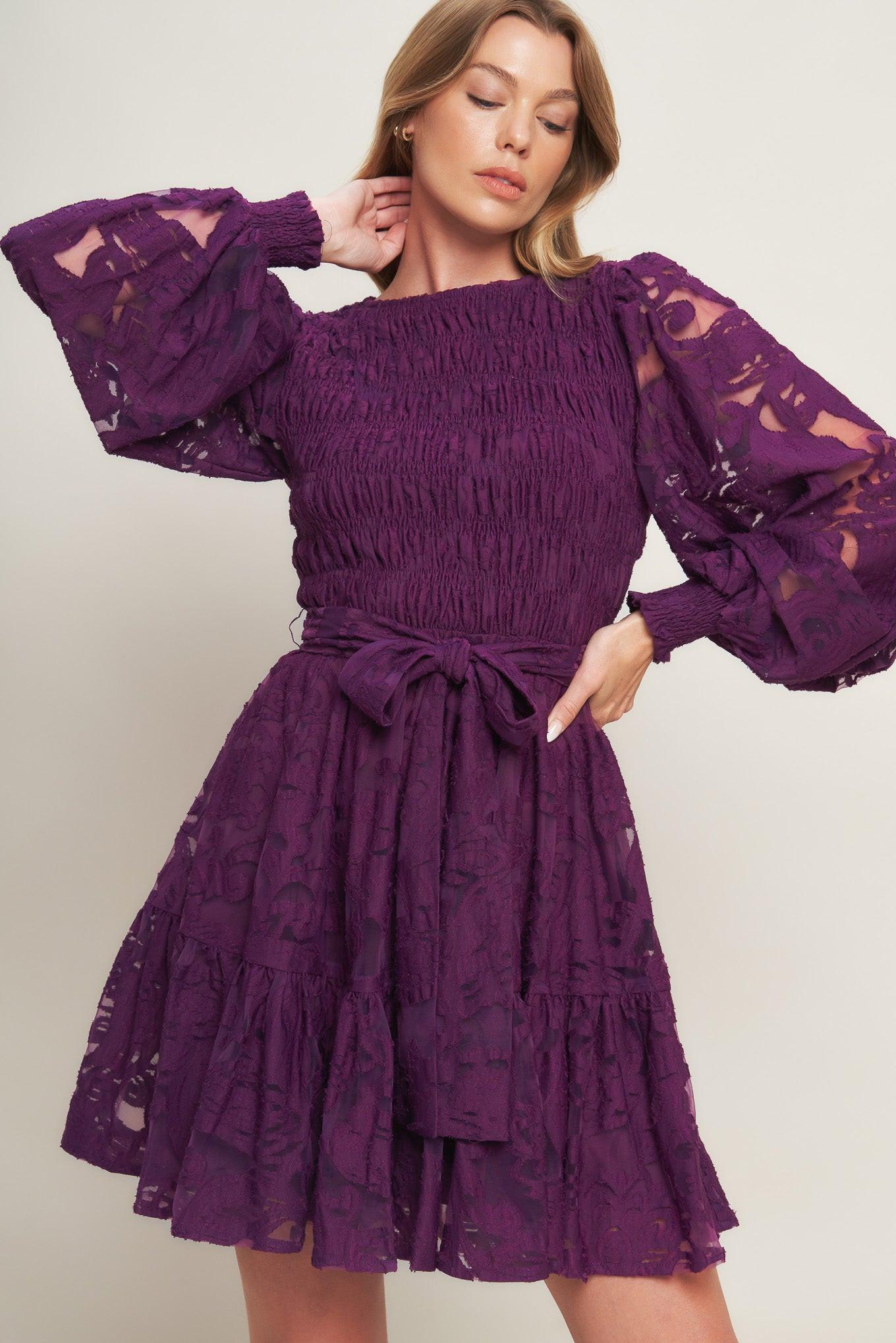 A woven lace mini dress featuring round neckline, long sleeve with cuff, smocked bodice, self sash tie, ruffled hemline and back neck button closure