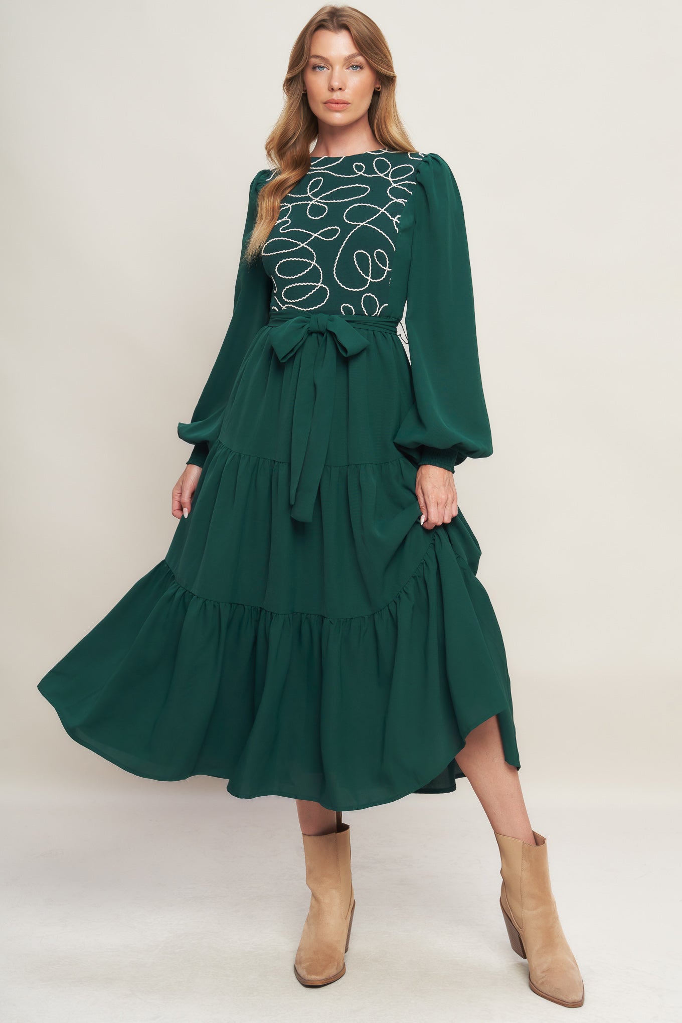 A solid woven midi dress featuring round neckline, long sleeve with smocked cuff, tiered skirt with self sash tie and back zipper closure