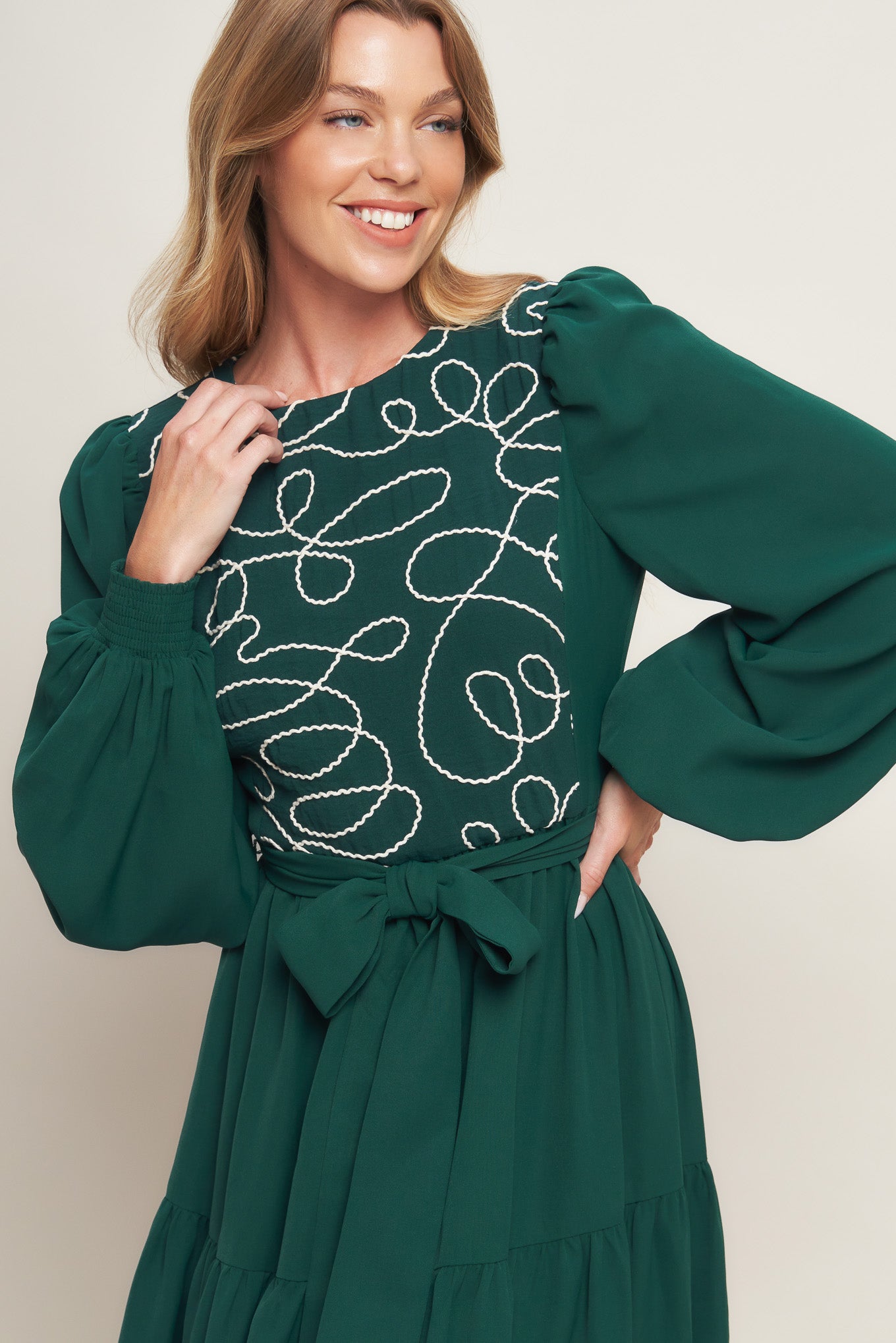 A solid woven midi dress featuring round neckline, long sleeve with smocked cuff, tiered skirt with self sash tie and back zipper closure