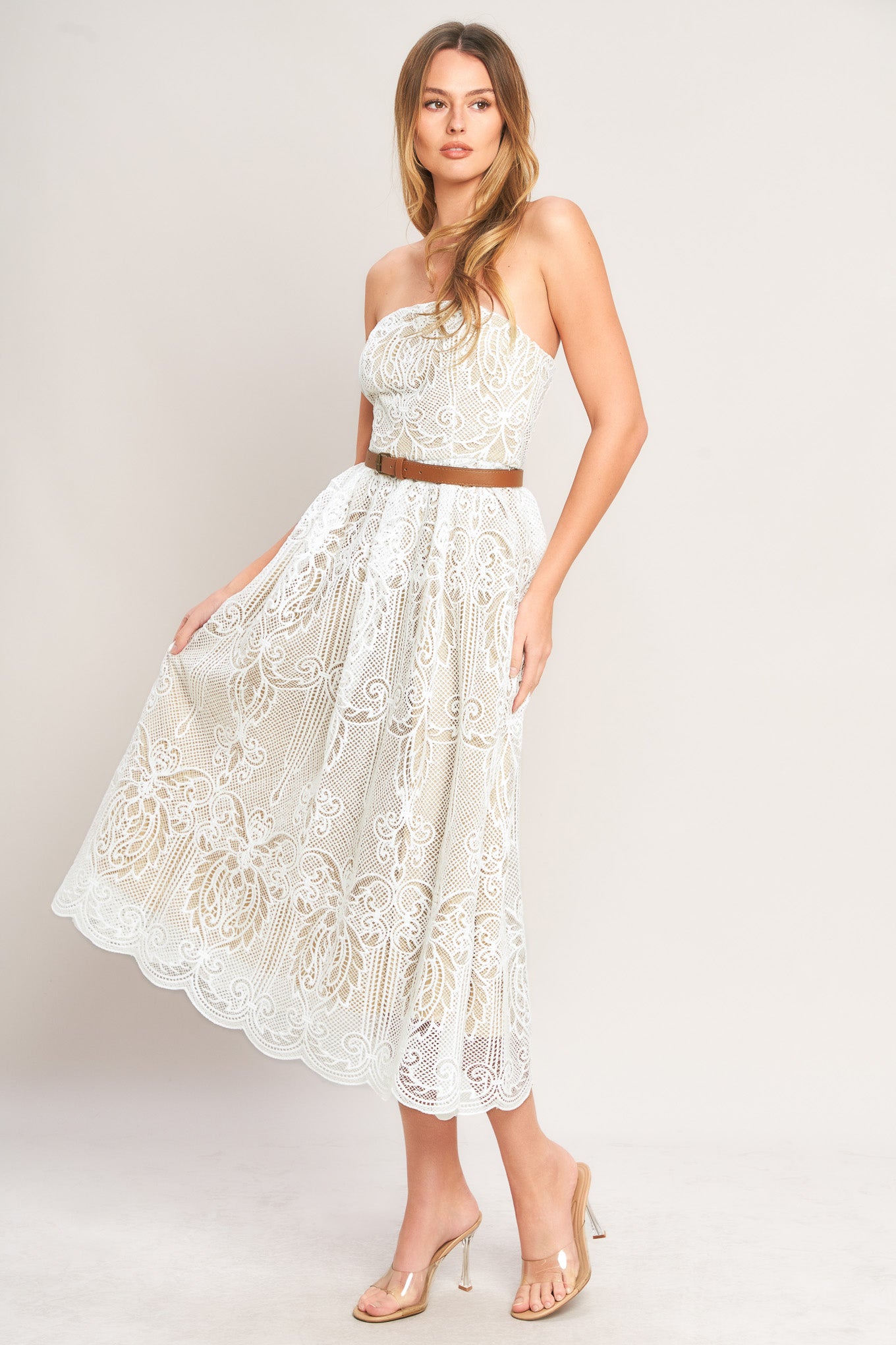 Woman wearing a white lace dress with a brown belt against a plain background